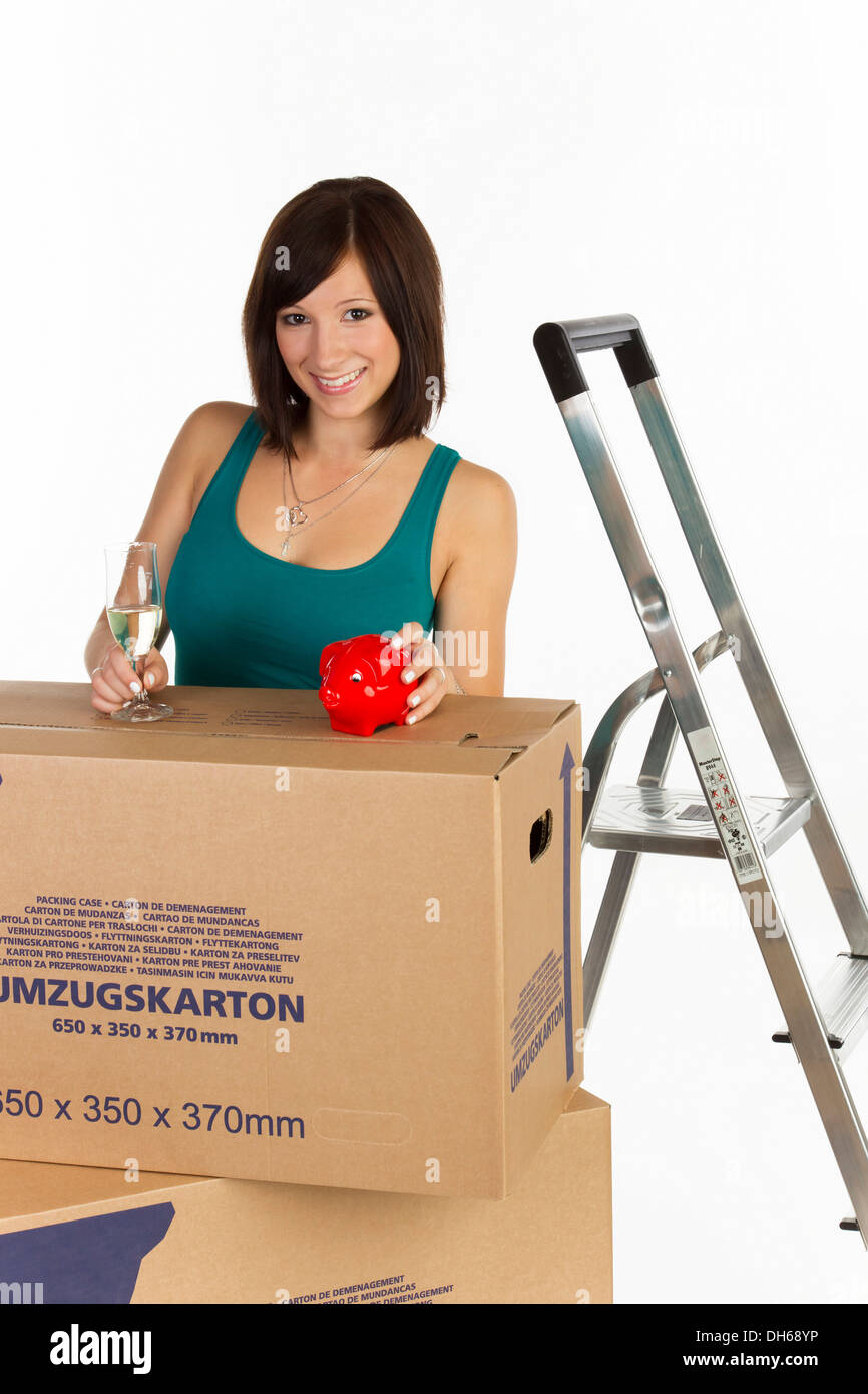 Young woman with moving boxes, a piggy bank and a glass of champagne in her hand Stock Photo