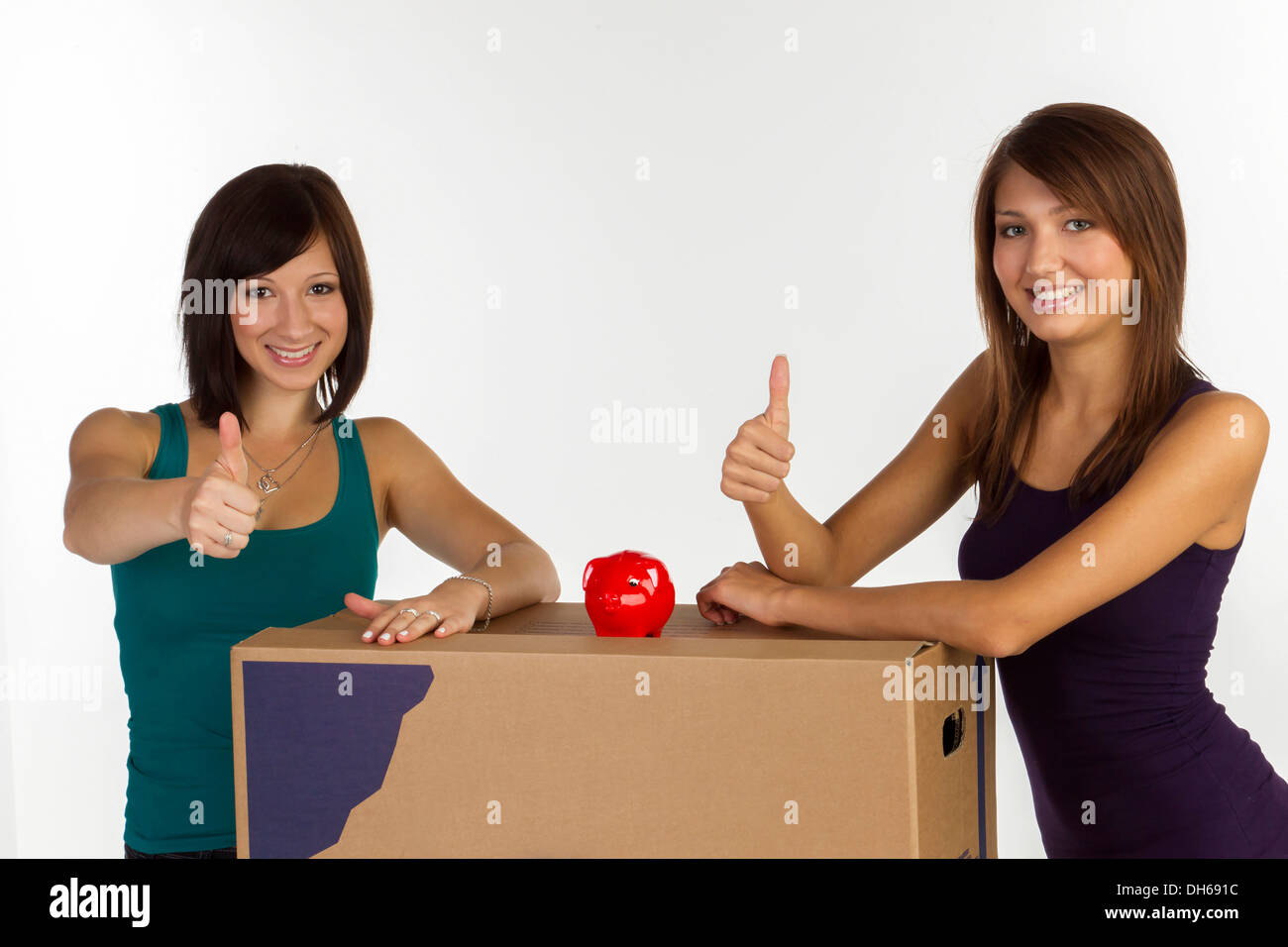 Two young woman with moving boxes and a piggy bank, making a thumbs-up gesture Stock Photo