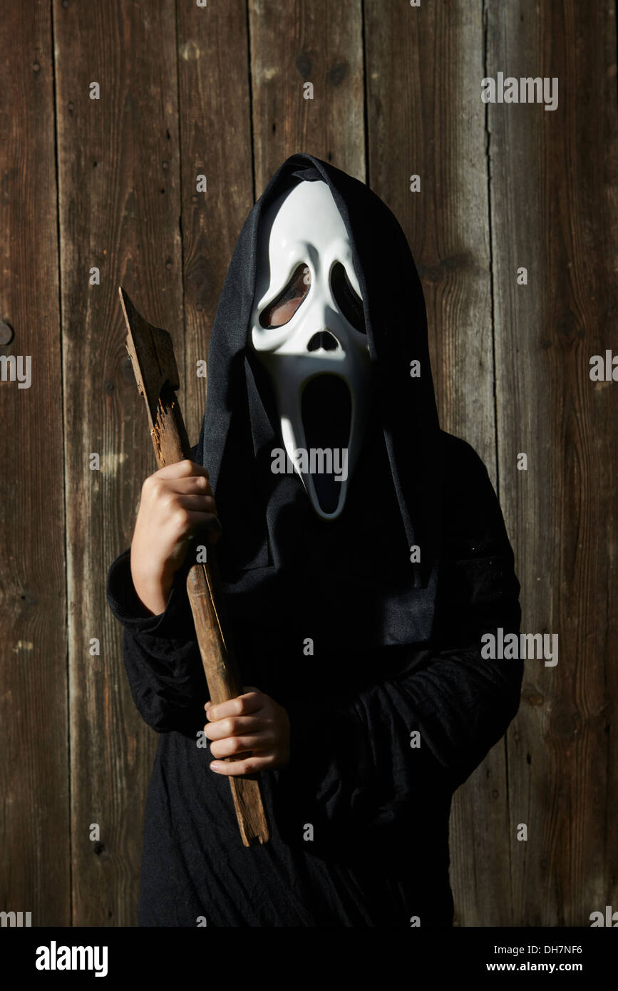 Man in scary 'scream' mask holds axe. Mask of grim reaper. Carnival white ghost mask and black hood. Scary Movie film. Stock Photo