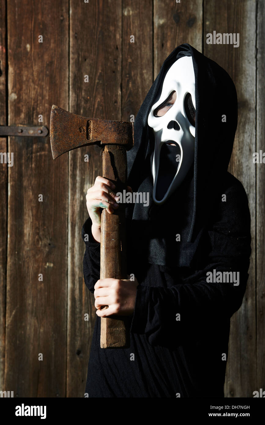 Man in scary 'scream' mask holds axe. Mask of grim reaper. Carnival white ghost mask and black hood. Scary Movie film theme. Stock Photo