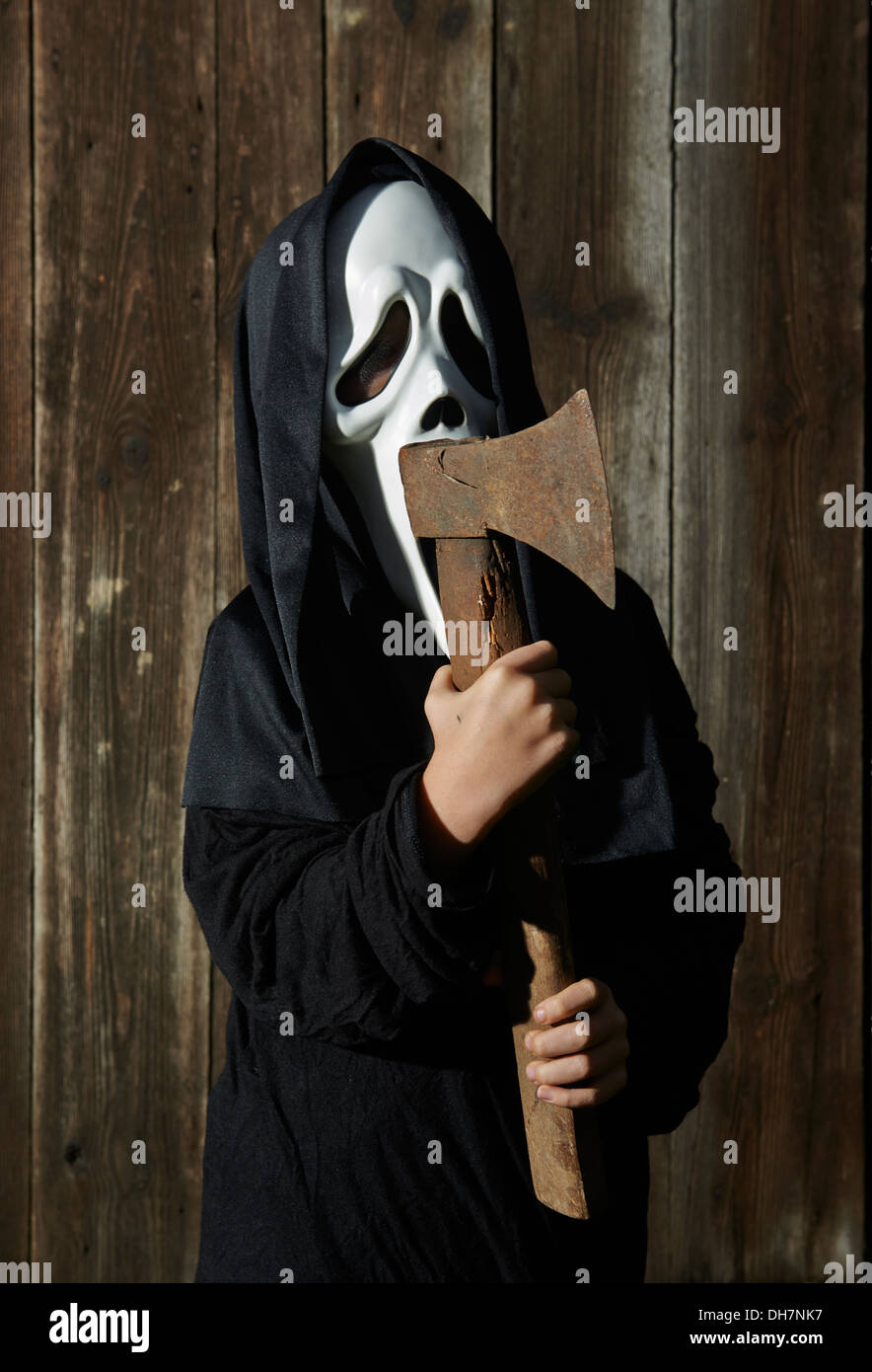 Man in scary 'scream' mask holds axe. Mask of grim reaper. Carnival white ghost mask and black hood. Scary Movie film. Stock Photo