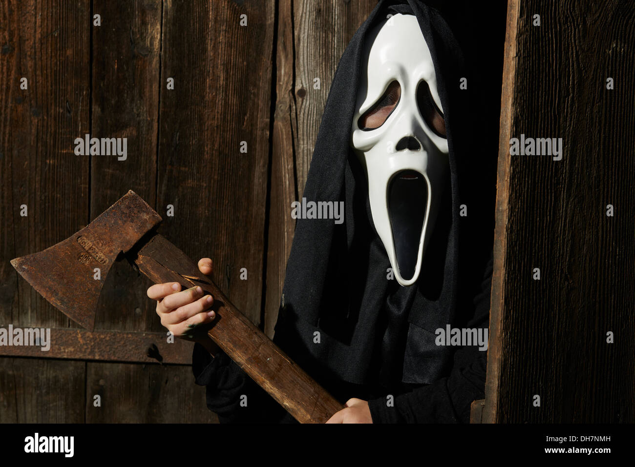 Man in scary 'scream' mask holds axe. Mask of grim reaper. Carnival white ghost mask and black hood. Scary Movie film. Stock Photo