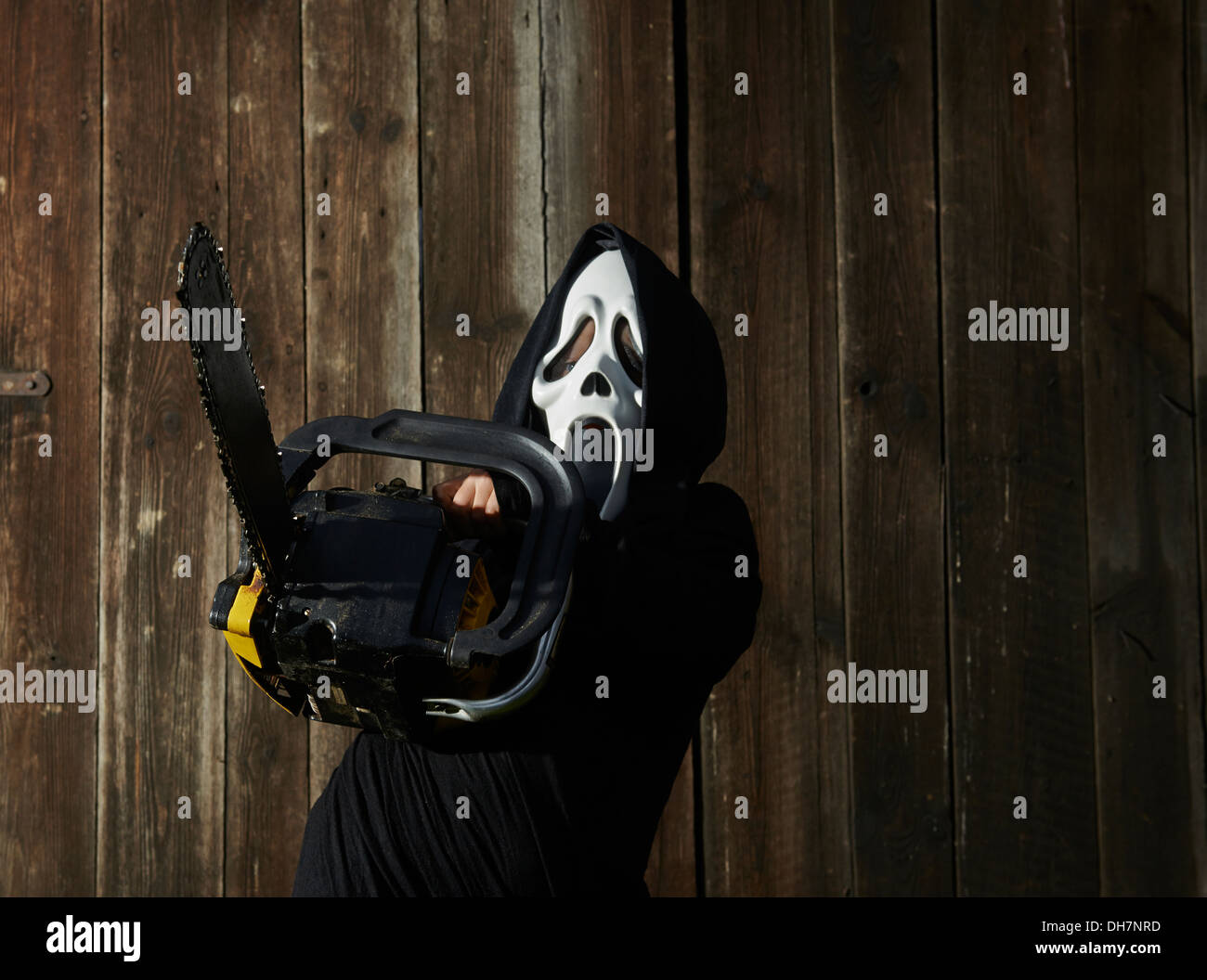 Man in scary 'scream' mask holds chainsaw. Mask of grim reaper. Carnival white ghost mask and black hood. Scary Movie film. Stock Photo