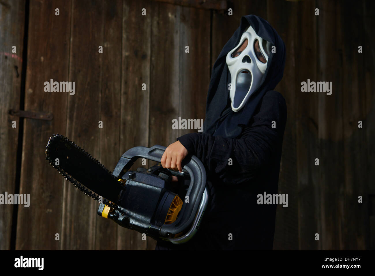 Man in scary 'scream' mask holds chainsaw. Mask of grim reaper. Carnival white ghost mask and black hood. Scary Movie film. Stock Photo