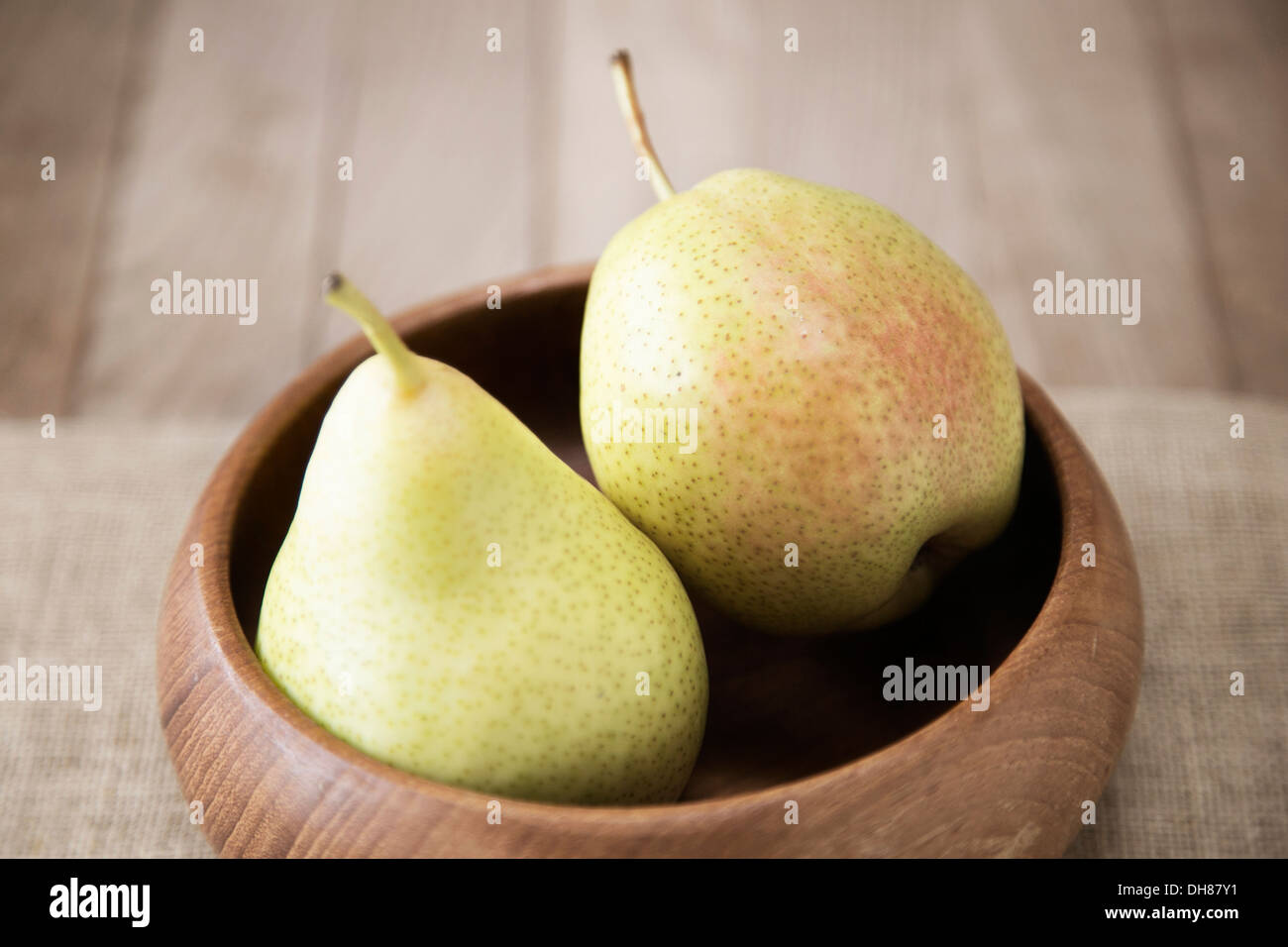 Two pears with mottled pale green skin tinged with pink in a wooden bowl with a linen cloth on a wooden surface Stock Photo