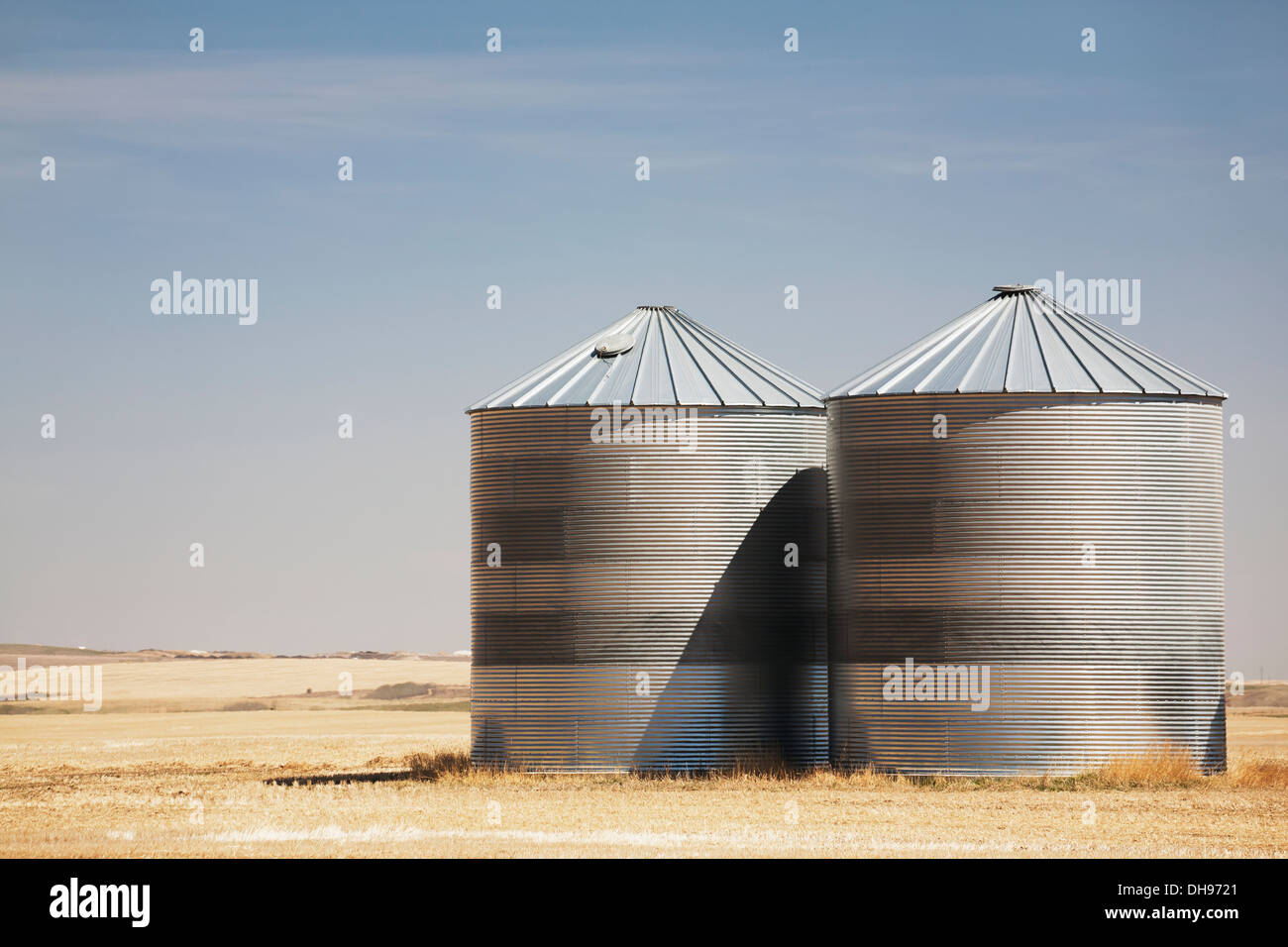 Two Large Grain Bins In A Cut Grain Field With Blue Sky And Clouds