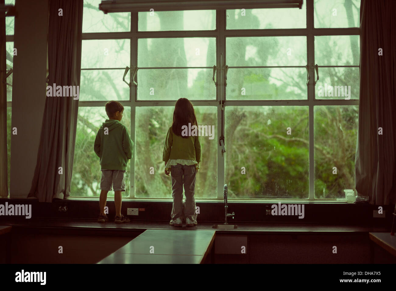 Children stand on bench and look out of large second floor window at trees. Stock Photo