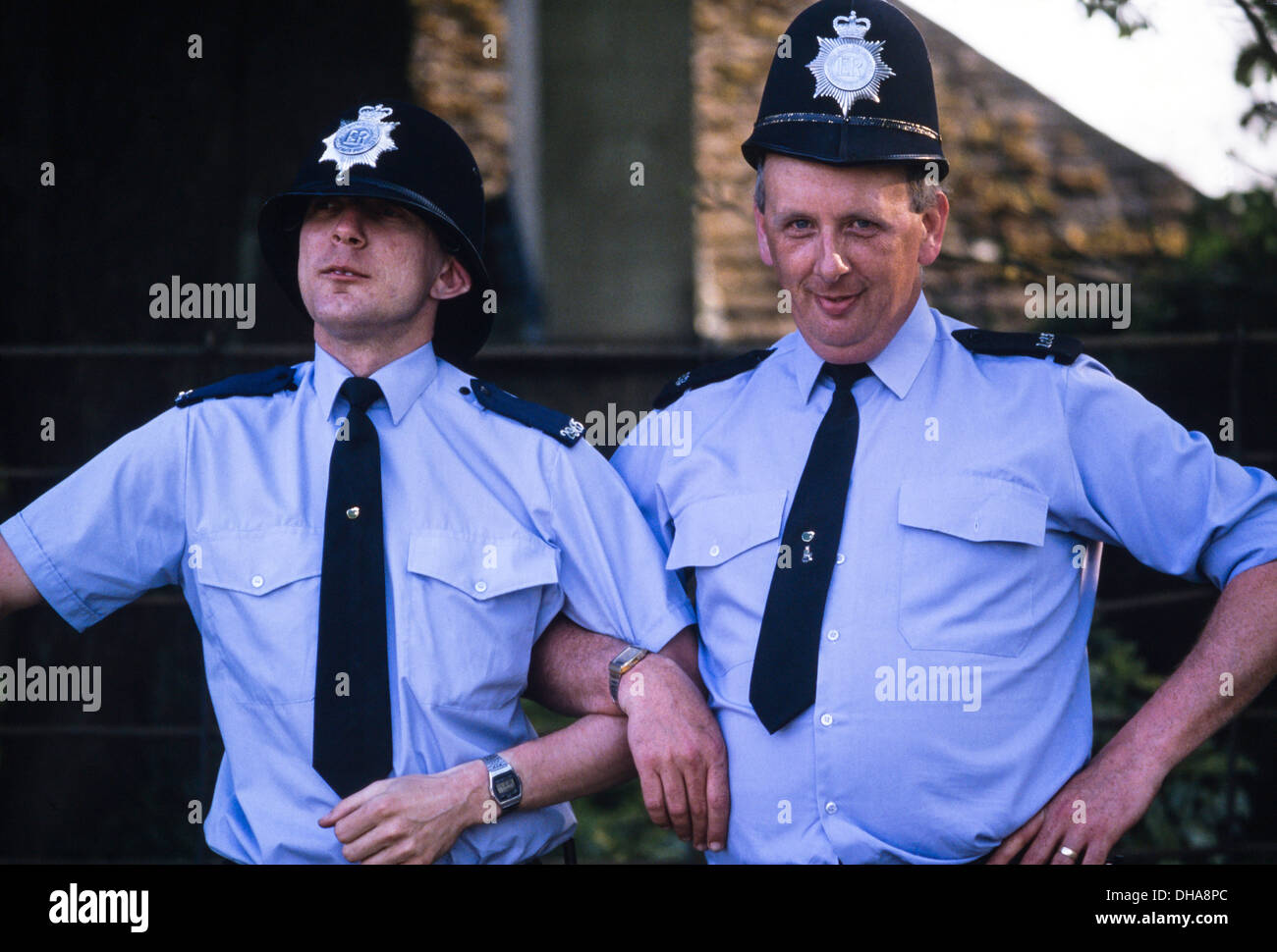 Two male police constables posing for a laugh with the wrong size custodian helmets. England, UK. Circa 1980's Stock Photo