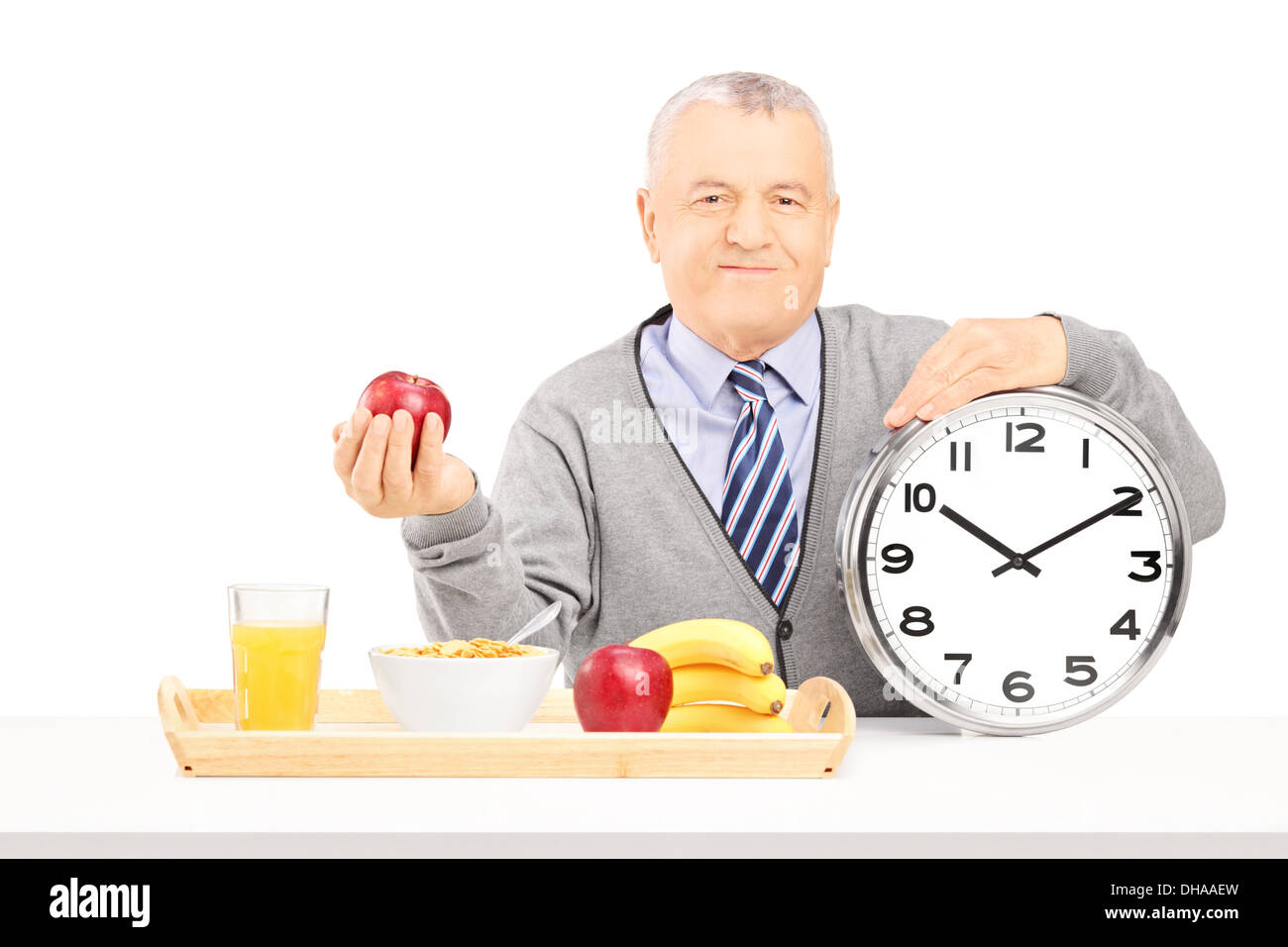 Senior gentleman holding a clock and red apple, on a table with a tray full of drink and food Stock Photo