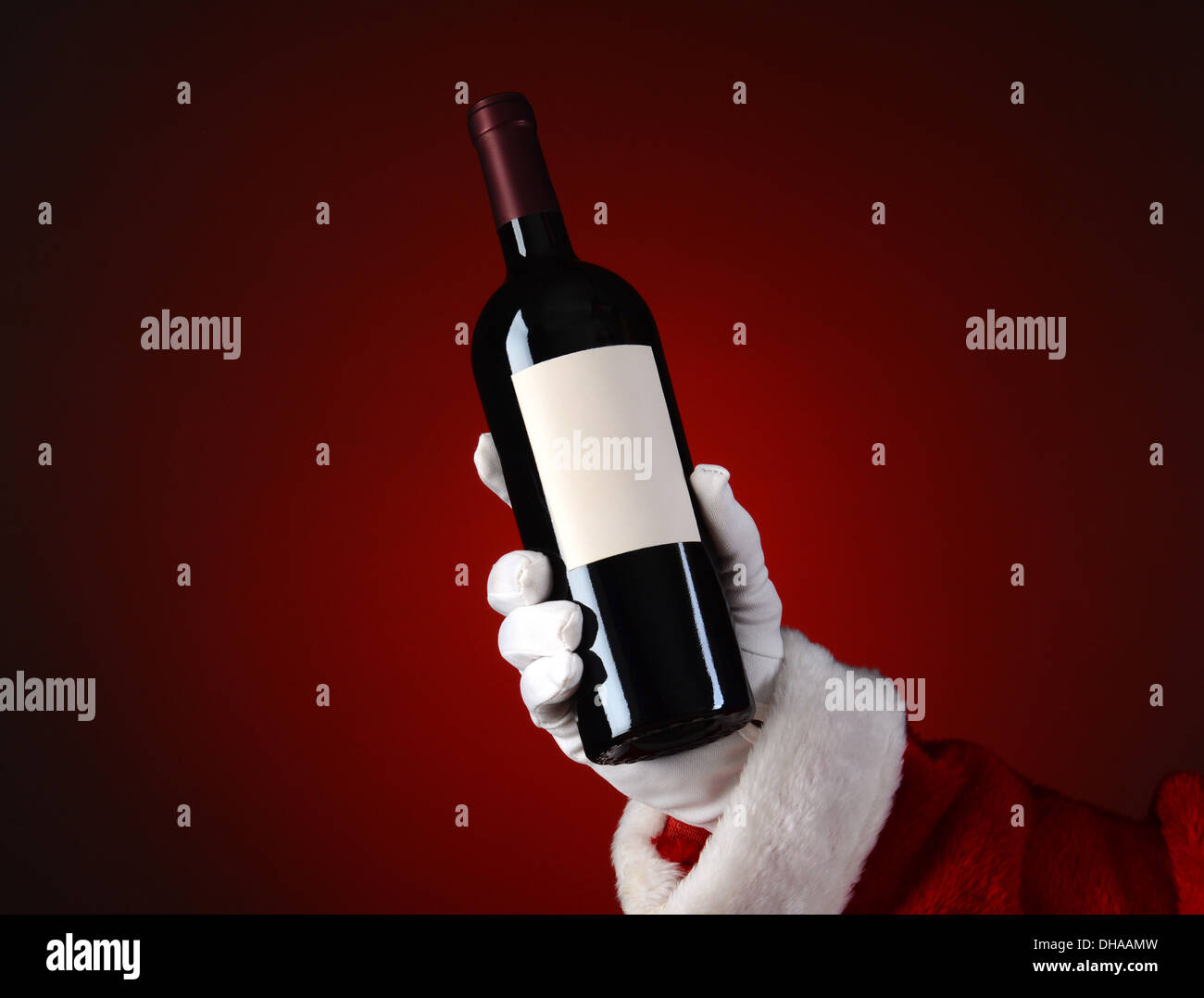 Closeup of Santa Claus holding a bottle of wine in his hand. Hand and arm only over a light to dark red spot background. Stock Photo