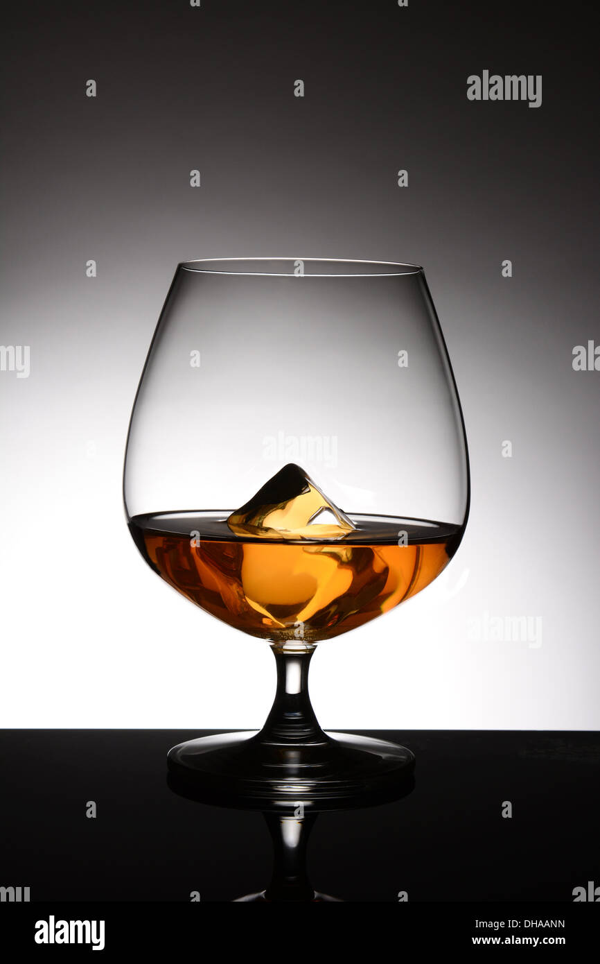 A brandy snifter with a single ice cube over a light to dark gray spot