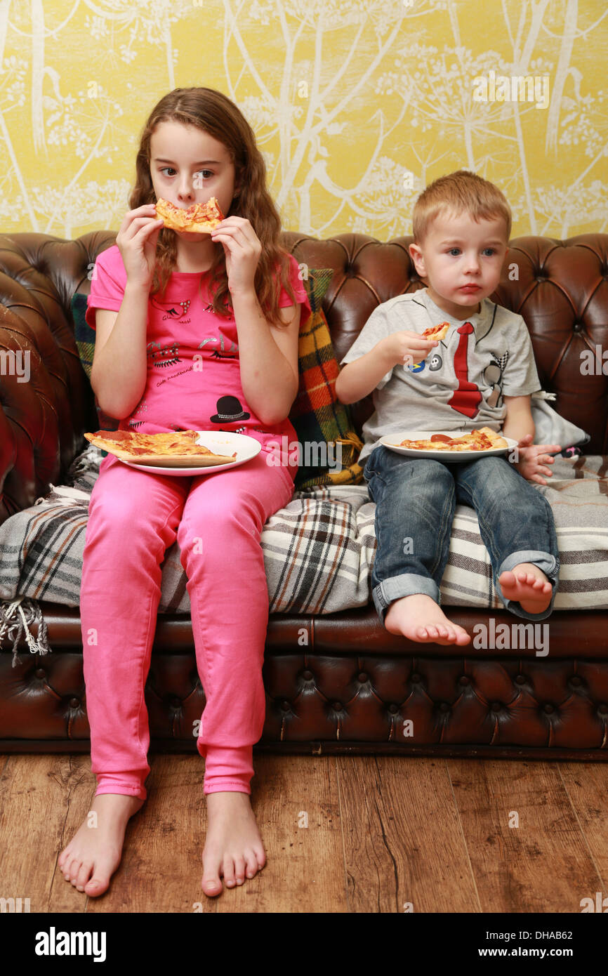 Brother and sister sitting on the sofa eating pizza while watching telly. Stock Photo