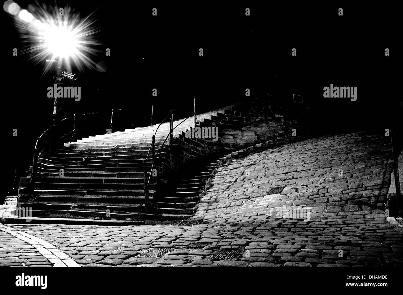 Black and white, low angle shot of cobbled street with stone staircase at left and traditional street lamp providing starburst. Stock Photo