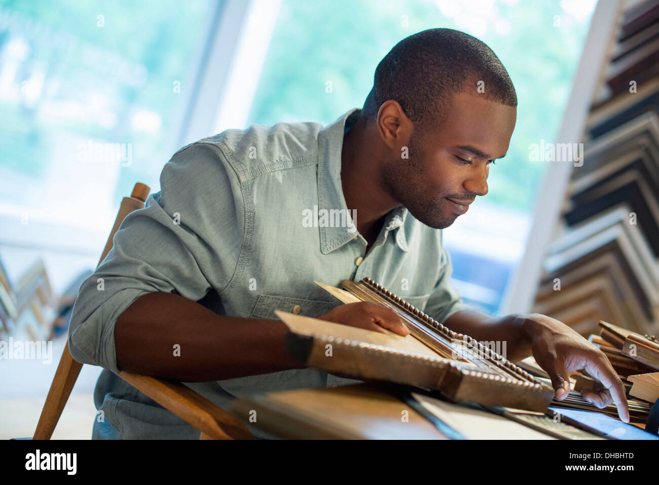 A young man at his workbench in a picture framing studio. Surrounded by samples. Using a digital tablet. Stock Photo