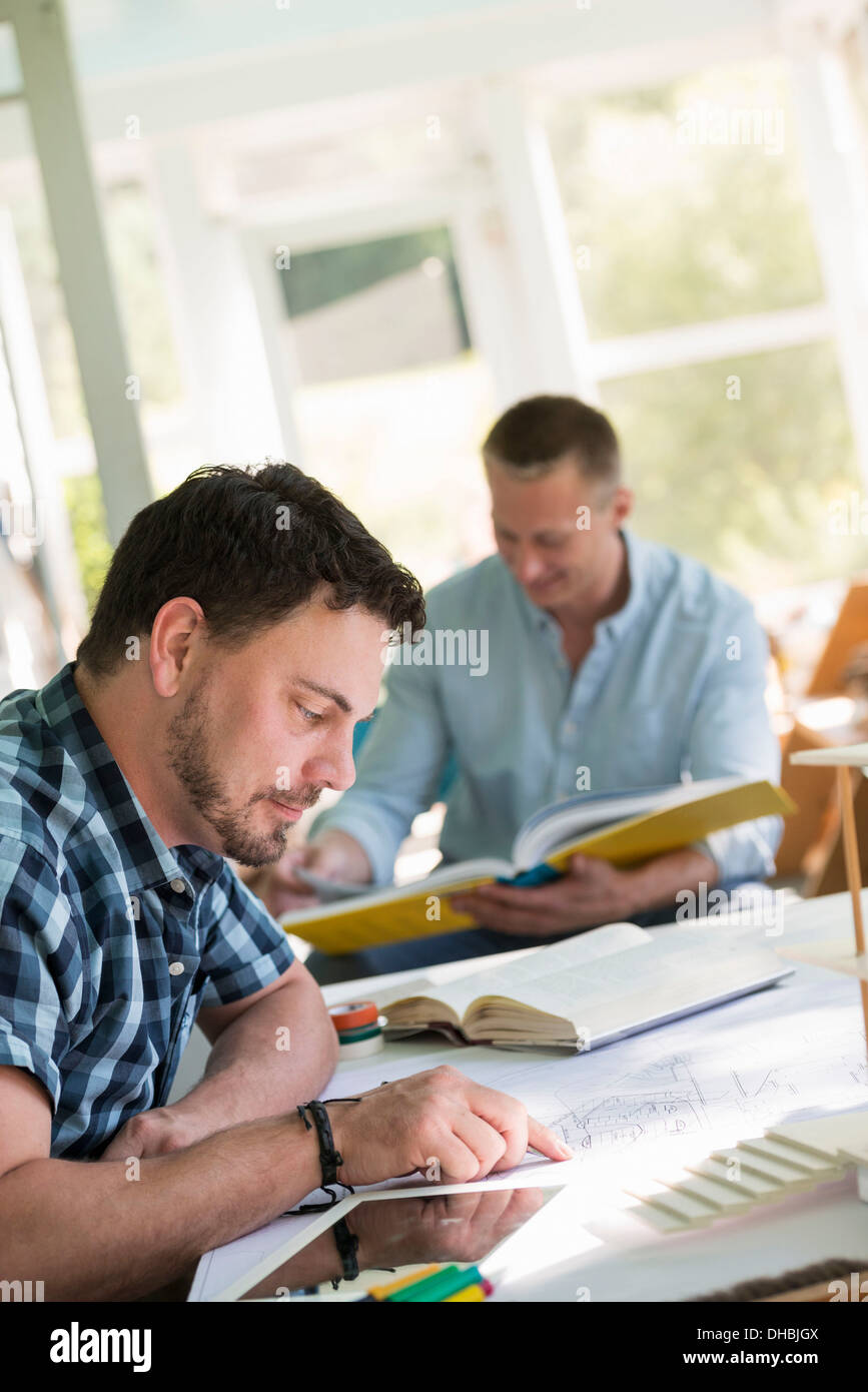 Two men seated at a table using books and a digital tablet. Stock Photo