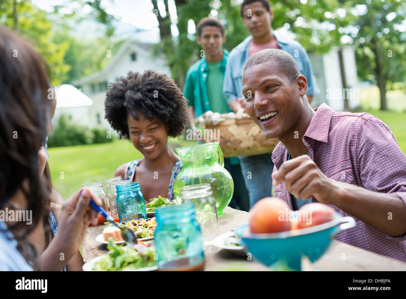 A group of adults and young people at a meal in the garden of a farmhouse. Passing plates and raising glasses. Stock Photo