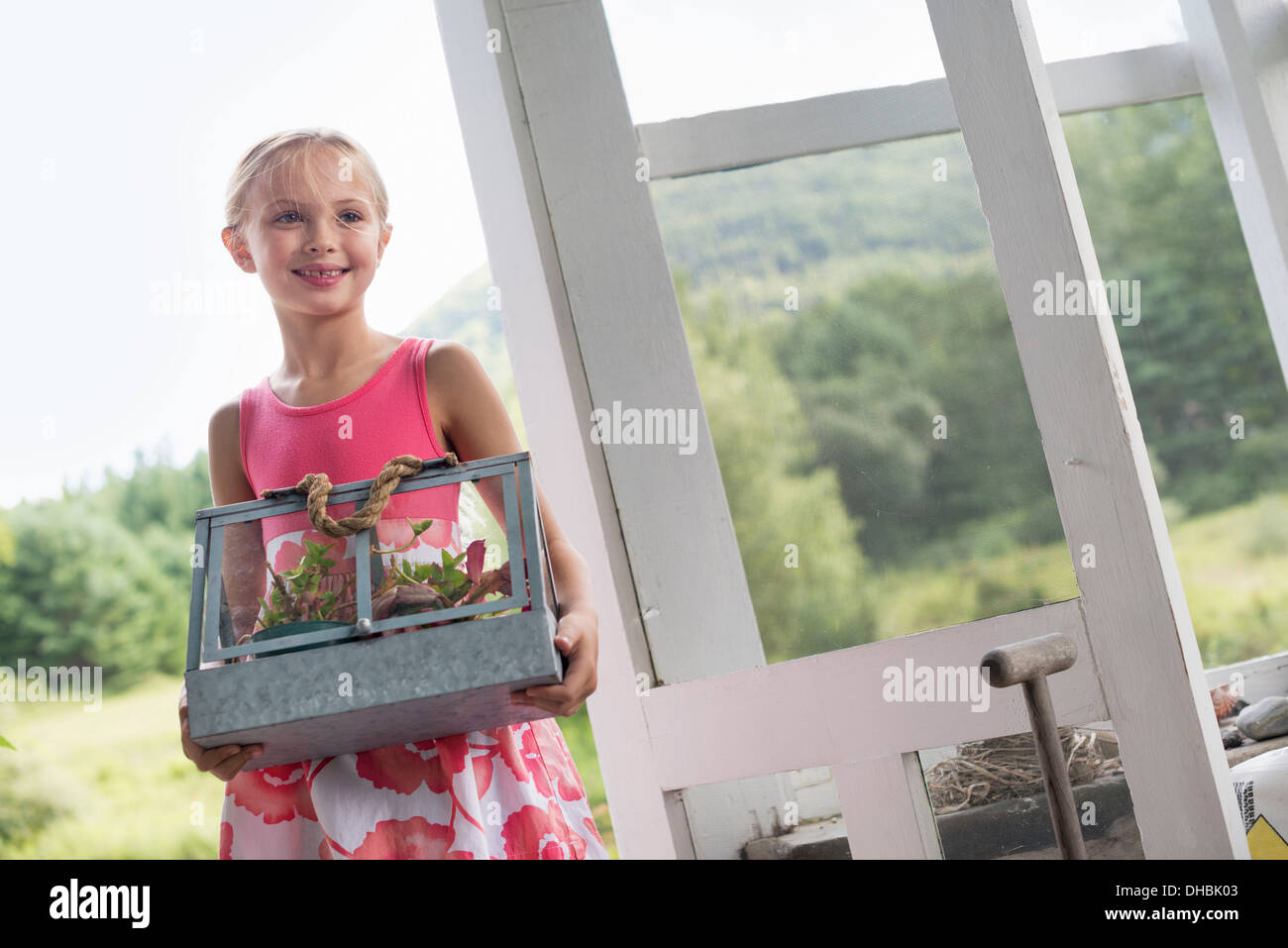 A young girl in a kitchen wearing a pink dress.  Carrying a terrarium containing small plants. Stock Photo