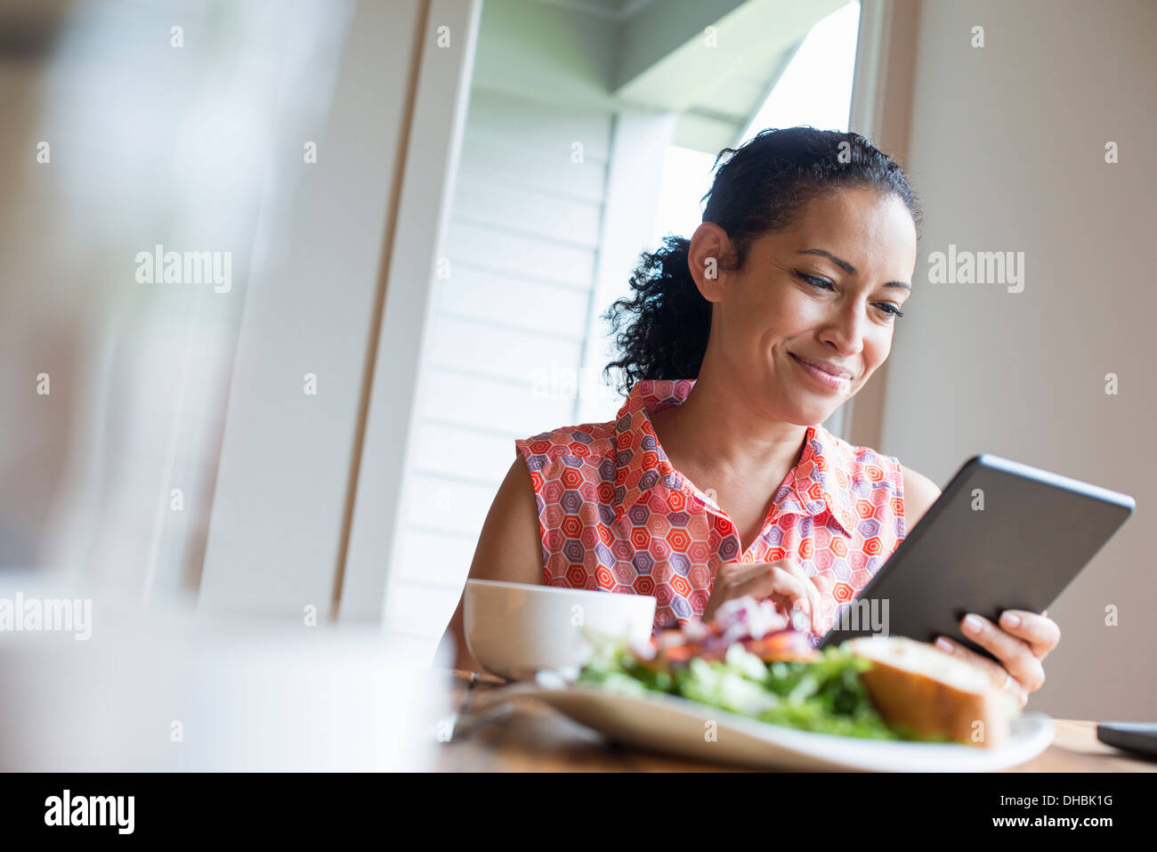 A young woman reading from the screen of a digital tablet, seated at a table. Coffee and a sandwich. Stock Photo