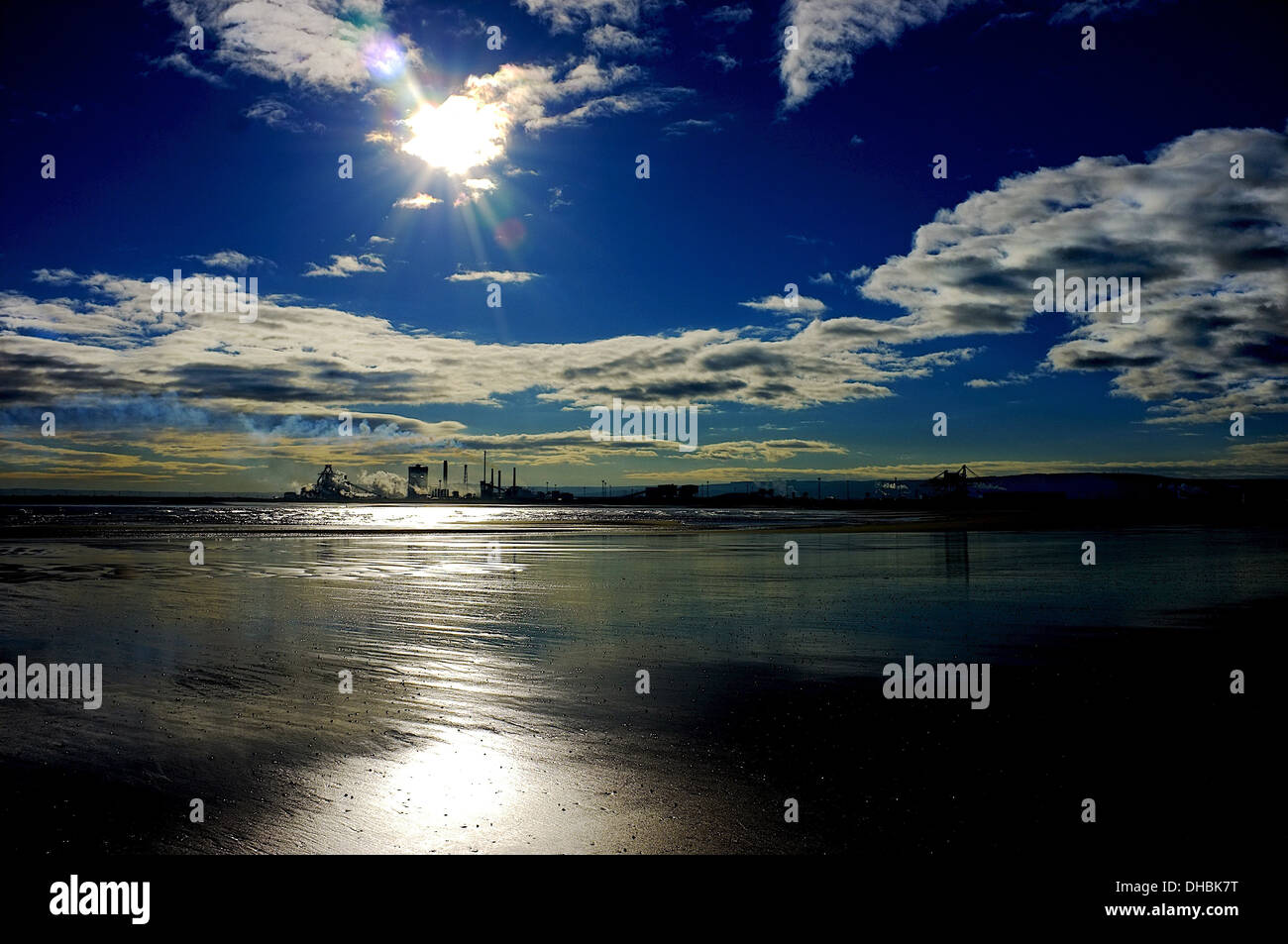 Early morning sun from a deep blue sky above a far off industrial installation creates a strip of light on a sea covered beach. Stock Photo