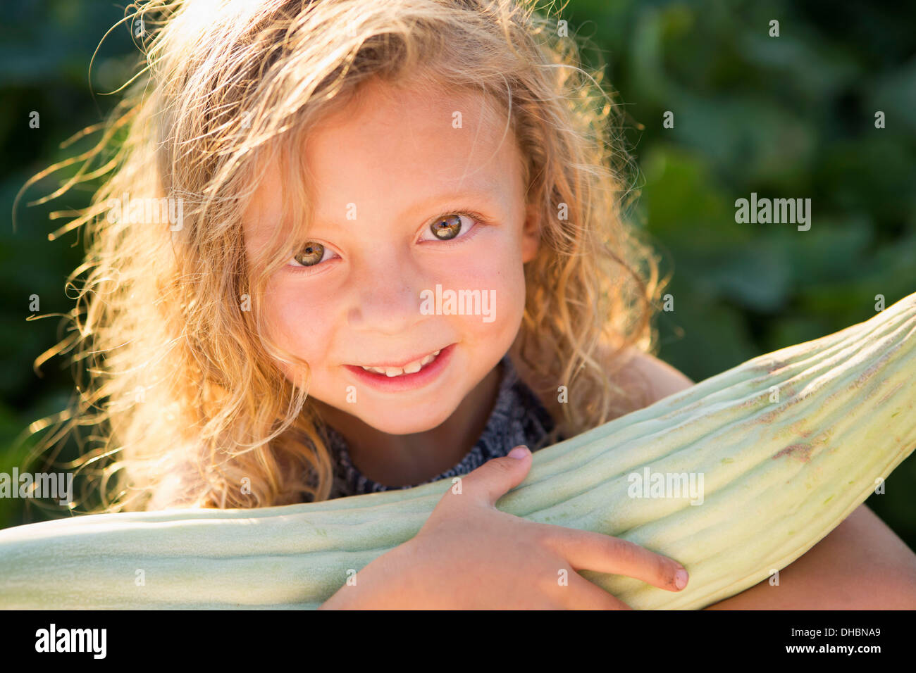 A young girl with long red curly hair outdoors in a garden holding a large fresh corn on the cob. Stock Photo