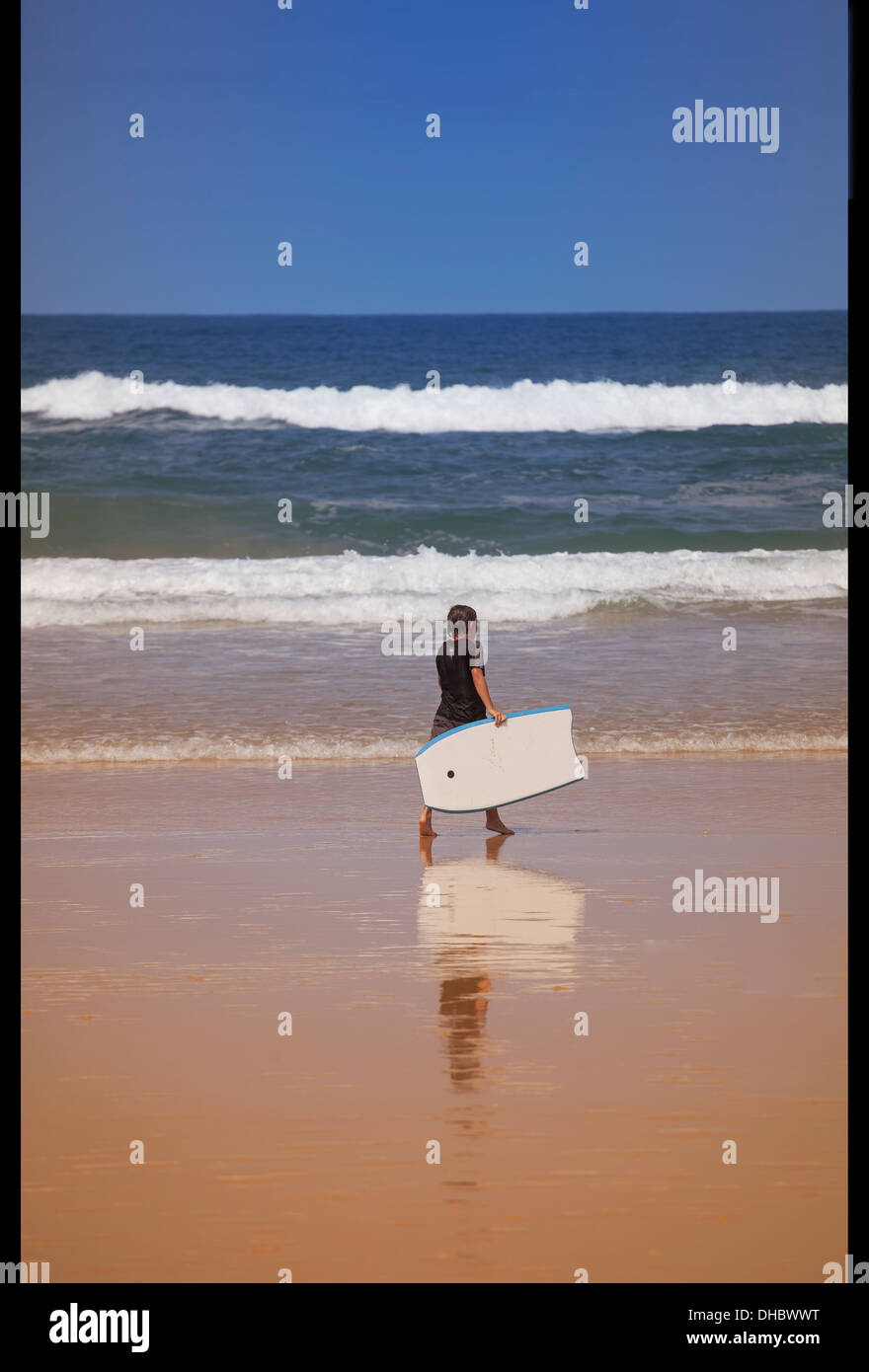 A white Australian male, teenager age 14-18, carrying a surf board on a beach in Australia. The image is in color. Stock Photo