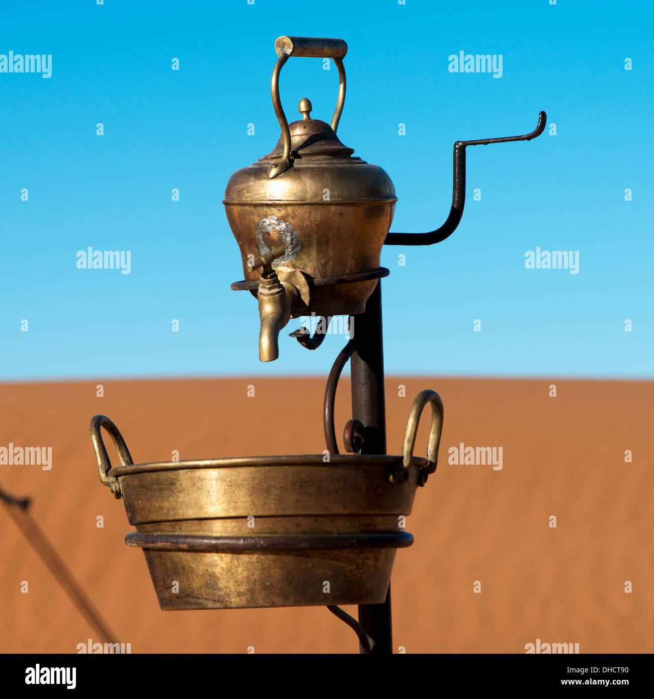 A Metal Teapot With A Tap And Bucket Below With A Sandy Landscape And Blue Sky Stock Photo