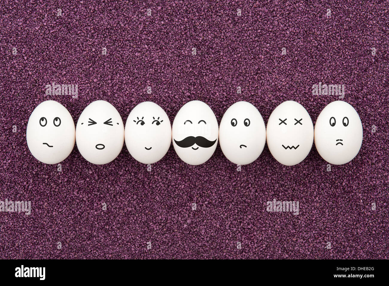 Seven eggs with different facial expressions are lying on the decorative purple sand. Stock Photo