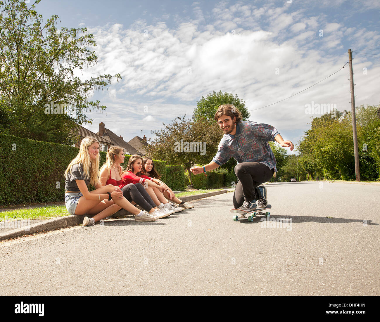Four young women sitting on kerb watching skateboarder Stock Photo