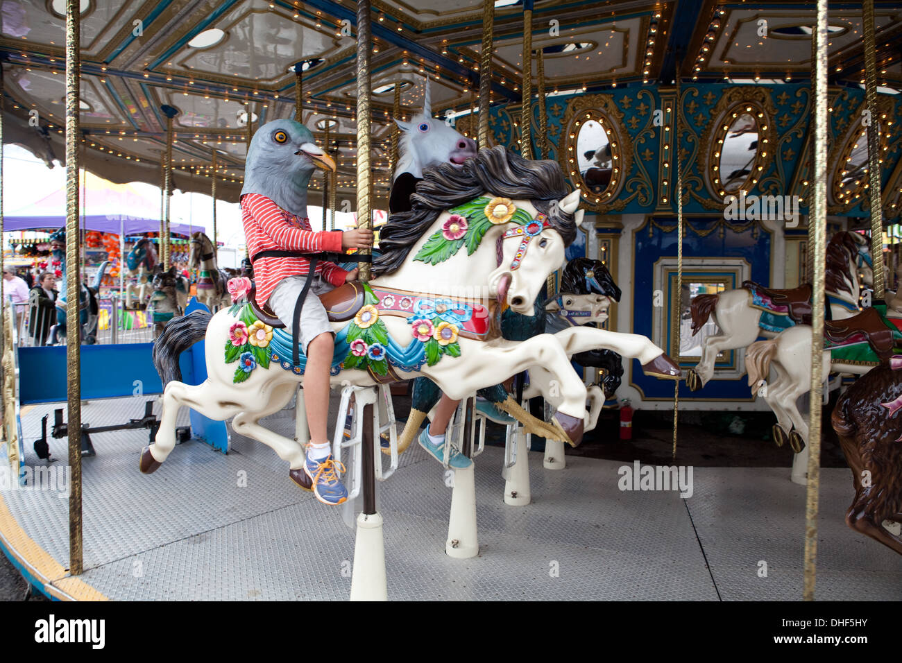 Girl wearing a pigeon mask rides horse on carousel. Stock Photo