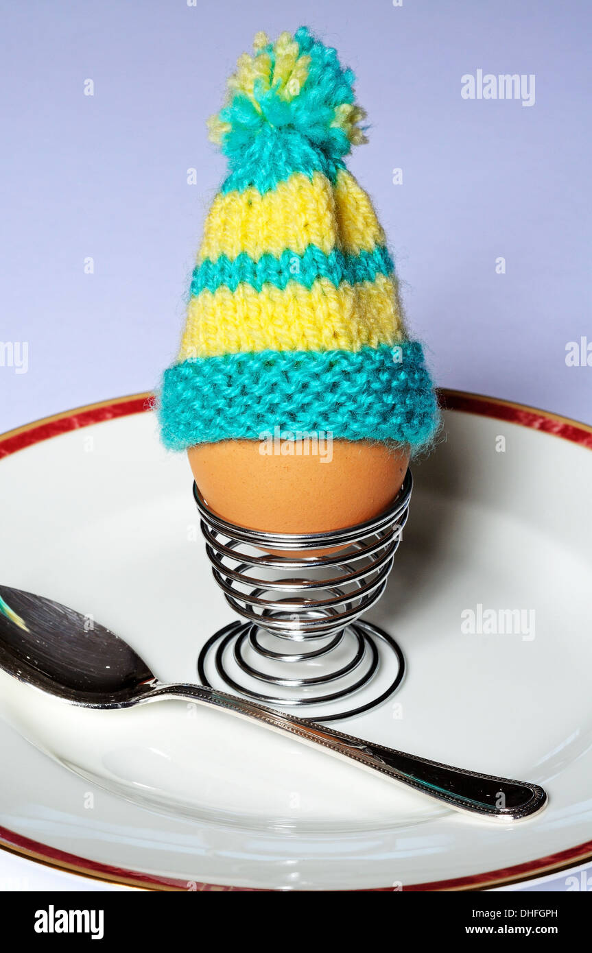 Boiled egg in a coiled metal eggcup topped with a knitted egg cosy on a white plate. Stock Photo