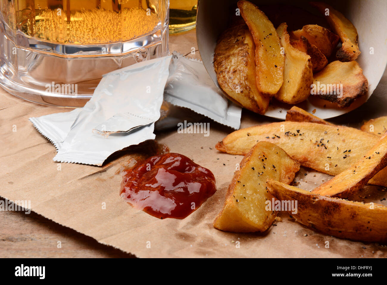French Fries spilled onto a brown bag. Ketchup dollop and packets with salt and pepper and mug of beer. Stock Photo