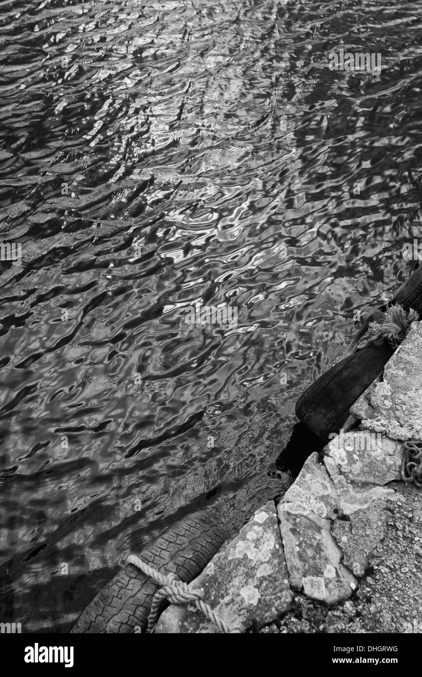 Black and white view of gently rippling water at edge of stone wall with two tyres attached with rope Stock Photo