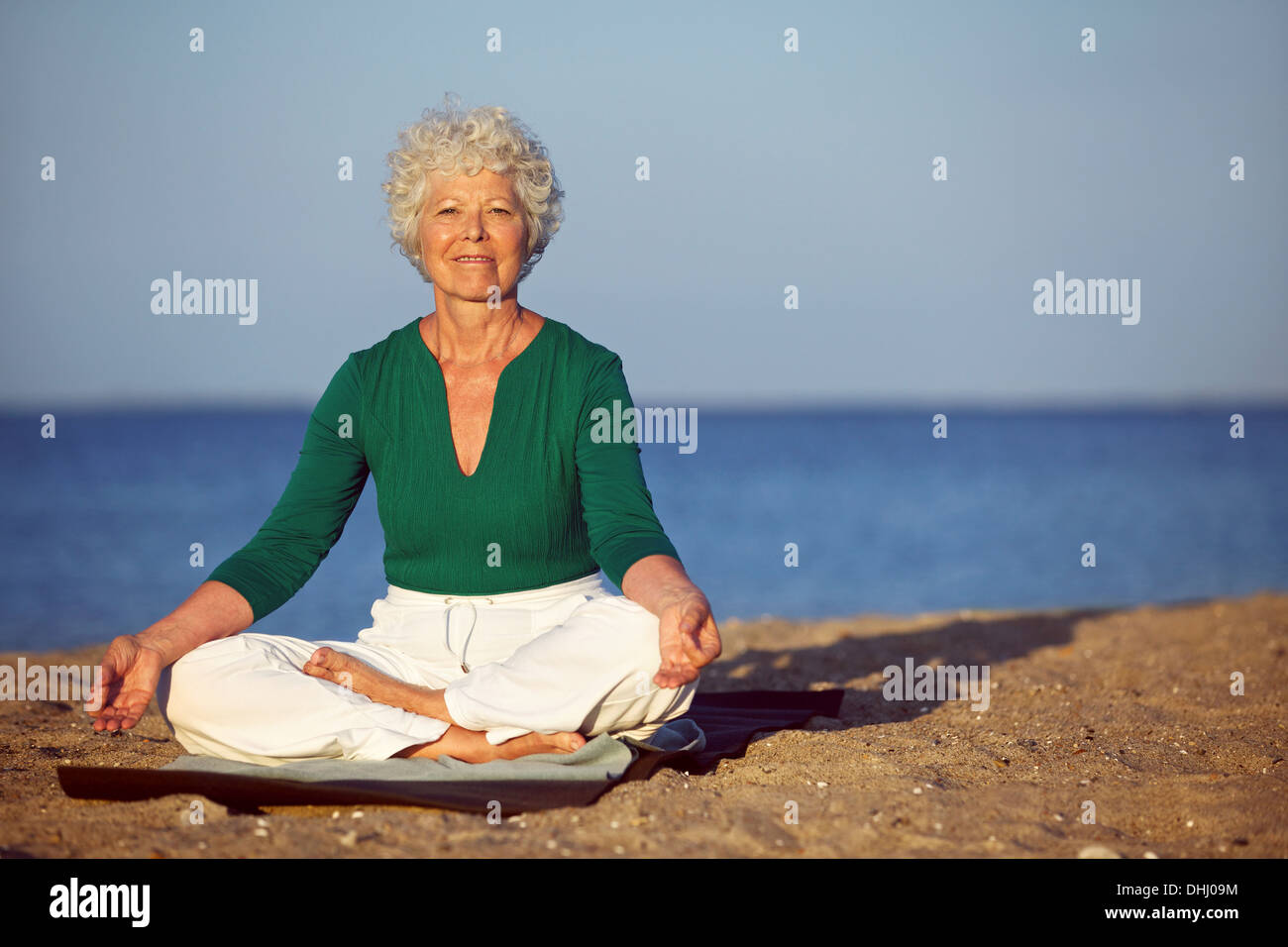 Portrait of senior woman meditating on sandy beach. Smiling mature woman exercising on seashore with copyspace. Stock Photo