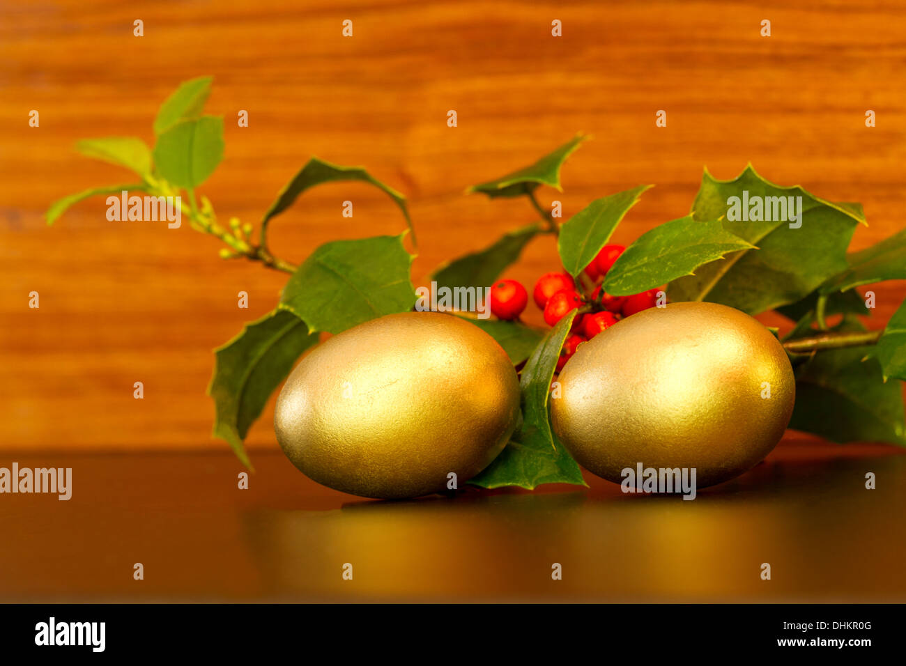Two gold nest eggs with festive green holly sprigs and red berries placed on wood grain background. Stock Photo