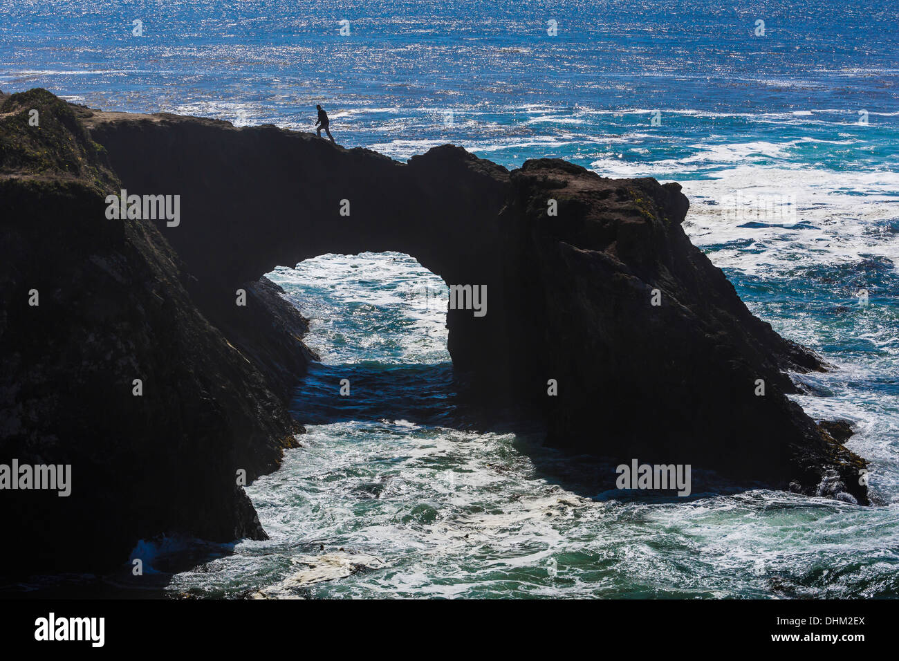 Man hiking atop arch with the roiling Pacific Ocean below, at Mendocino Headlands State Park near Mendocino, California, USA Stock Photo