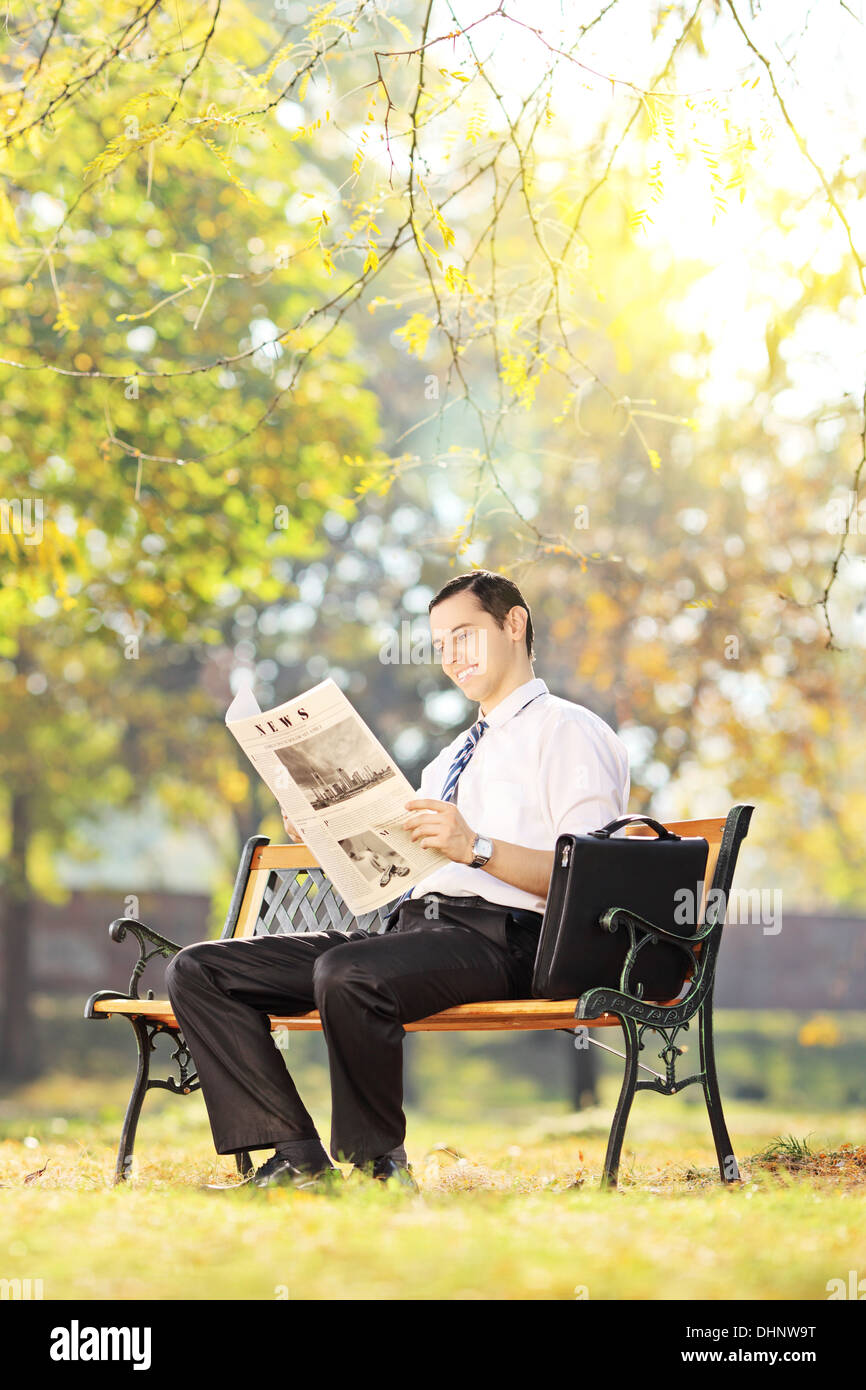 Young man seated on a bench reading a newspaper in a park on a sunny ...