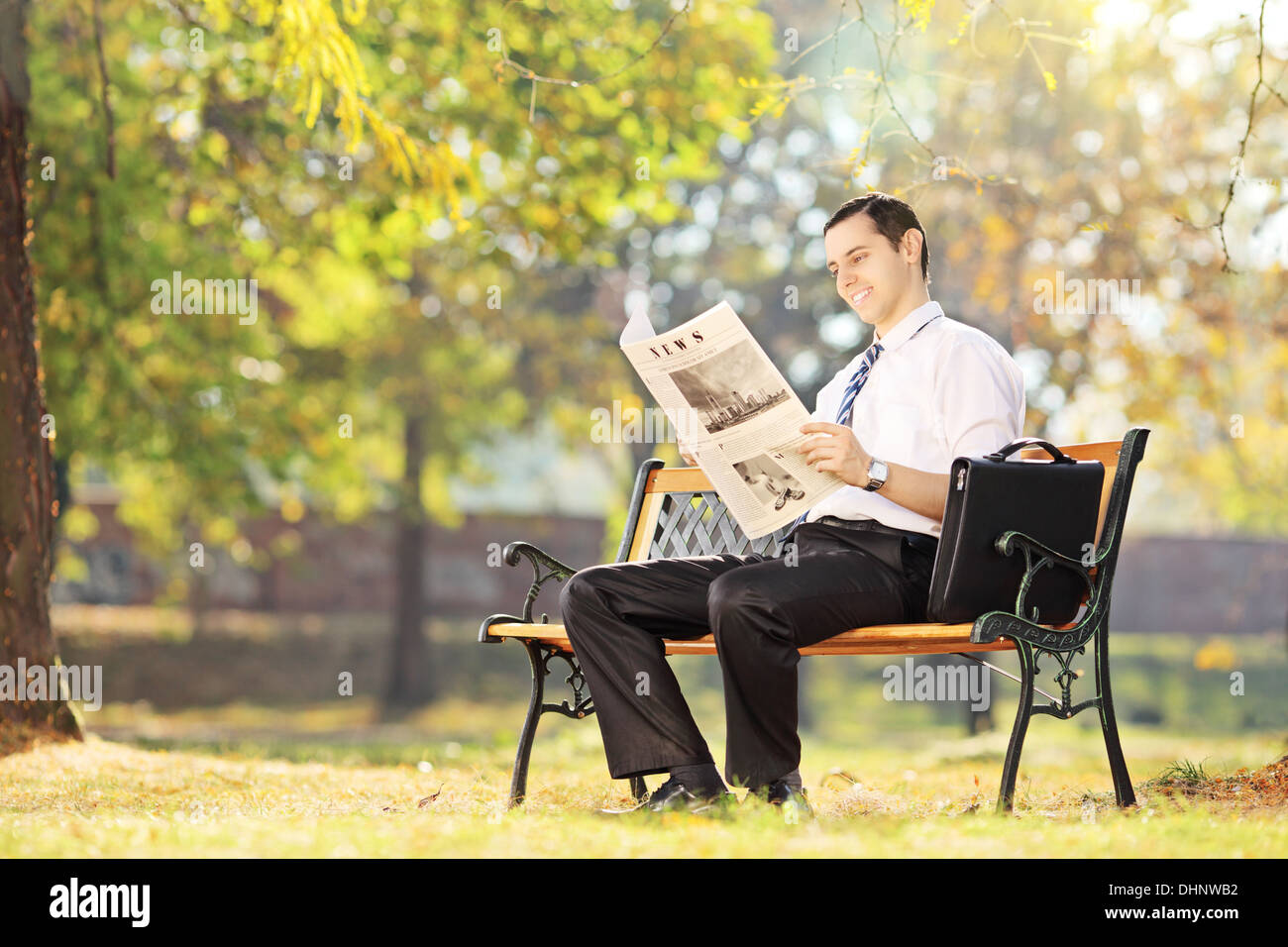 Young smiling man seated on a bench reading a newspaper in a park Stock ...