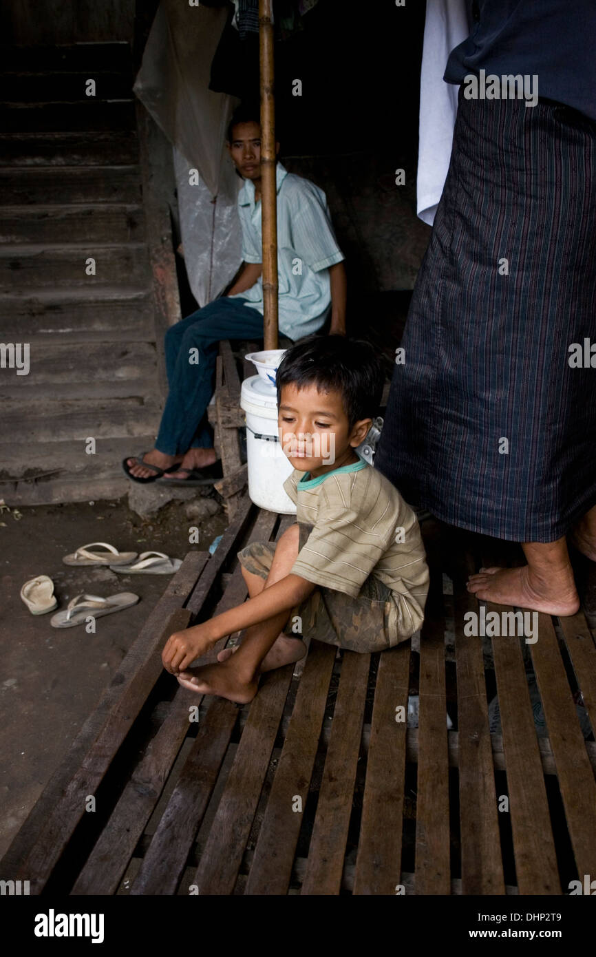 A young child laborer boy living in poverty is sitting on a wooden bed in a ramshackle hovel in Kampong Cham, Cambodia. Stock Photo