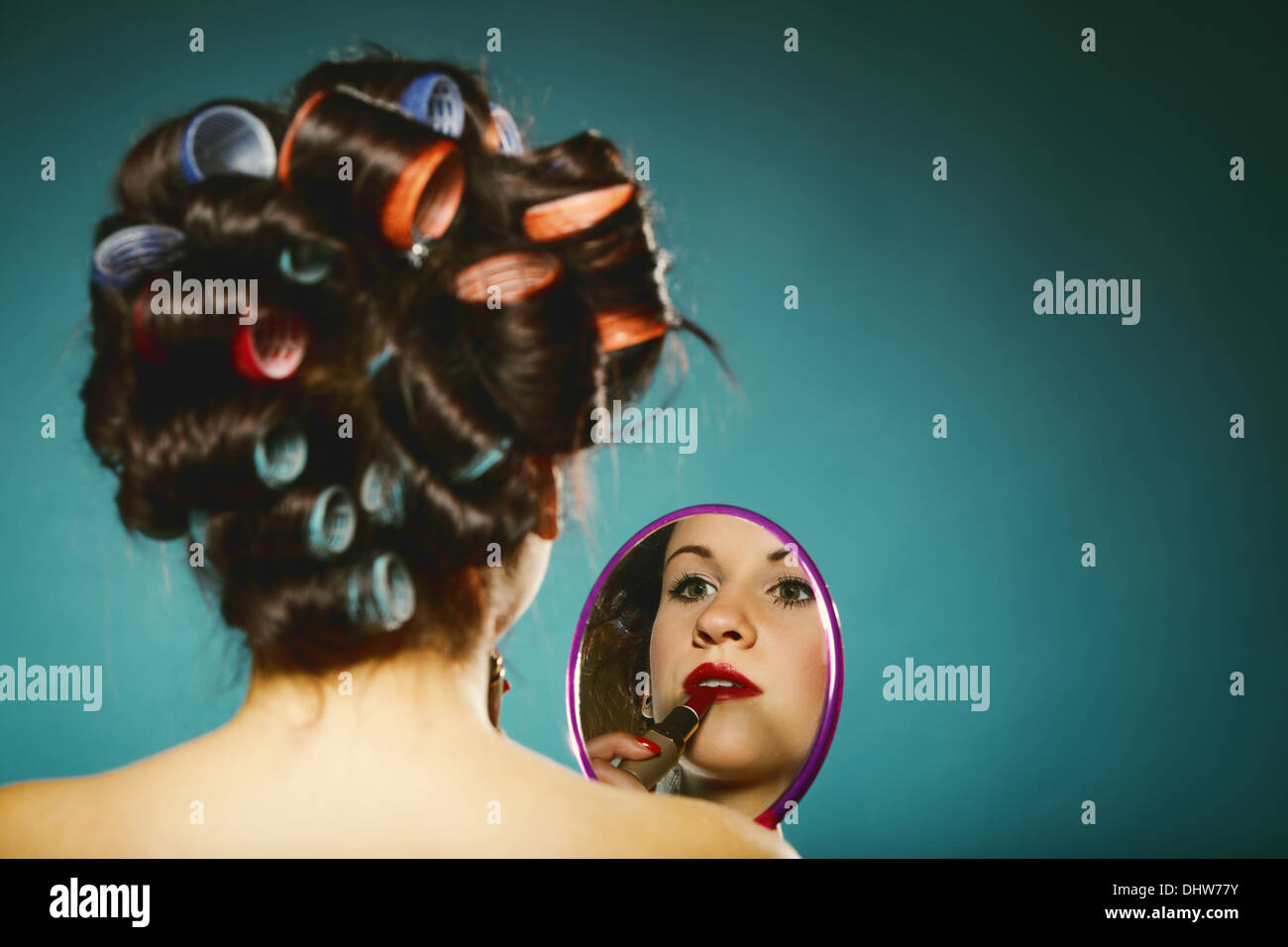 Young woman preparing to party having fun, girl styling hair with curlers looking in the mirror retro style blue background Stock Photo