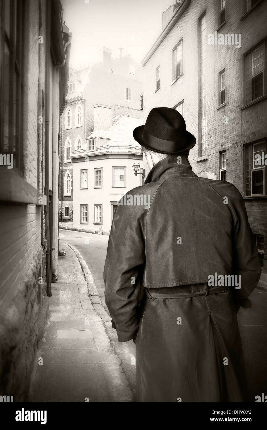 Black and white image of a man in a hat and trench coat from behind on old city street with pub in the distance. Stock Photo