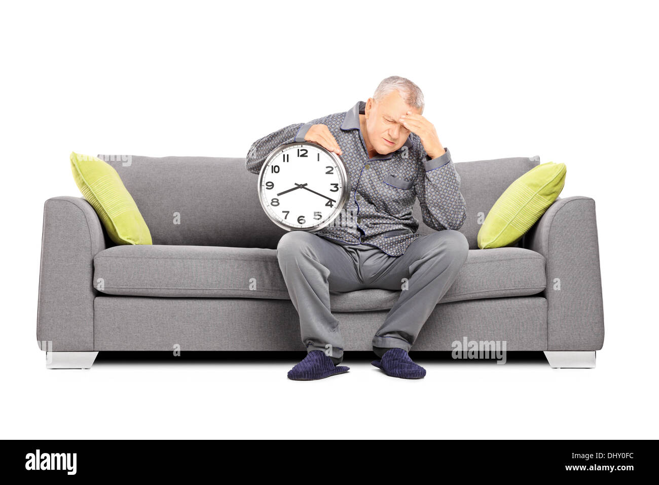 Mature man in pajamas seated on a sofa holding a clock and having a headache Stock Photo