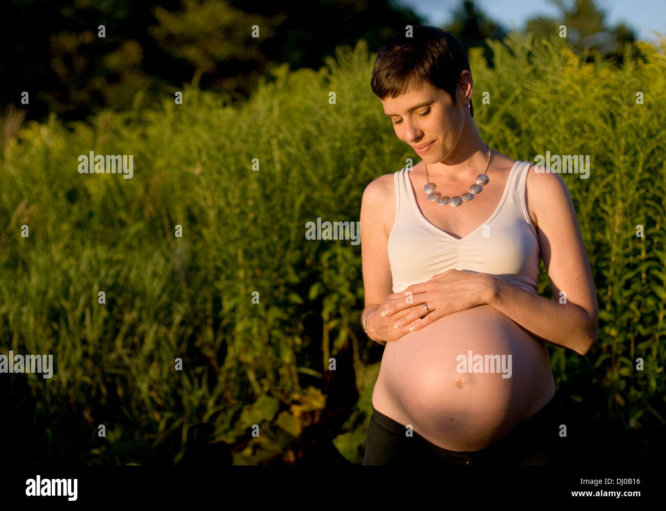 A nine-months pregnant Caucasian woman with short brown hair smiles softly in a field in late summer. Stock Photo