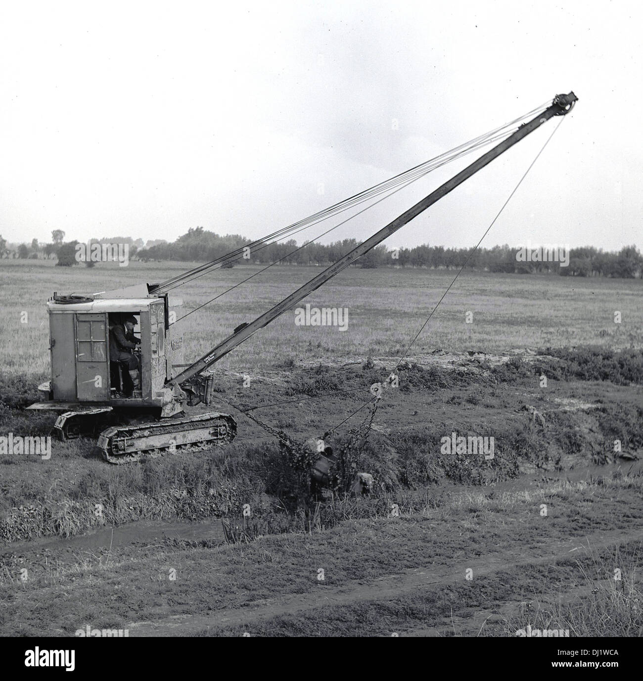 1950s, historical picture of a mechanical dragline bucket on crane with tracks positioned in a country field, excavating earth from a small river or brook that runs beside it, England, UK. Stock Photo