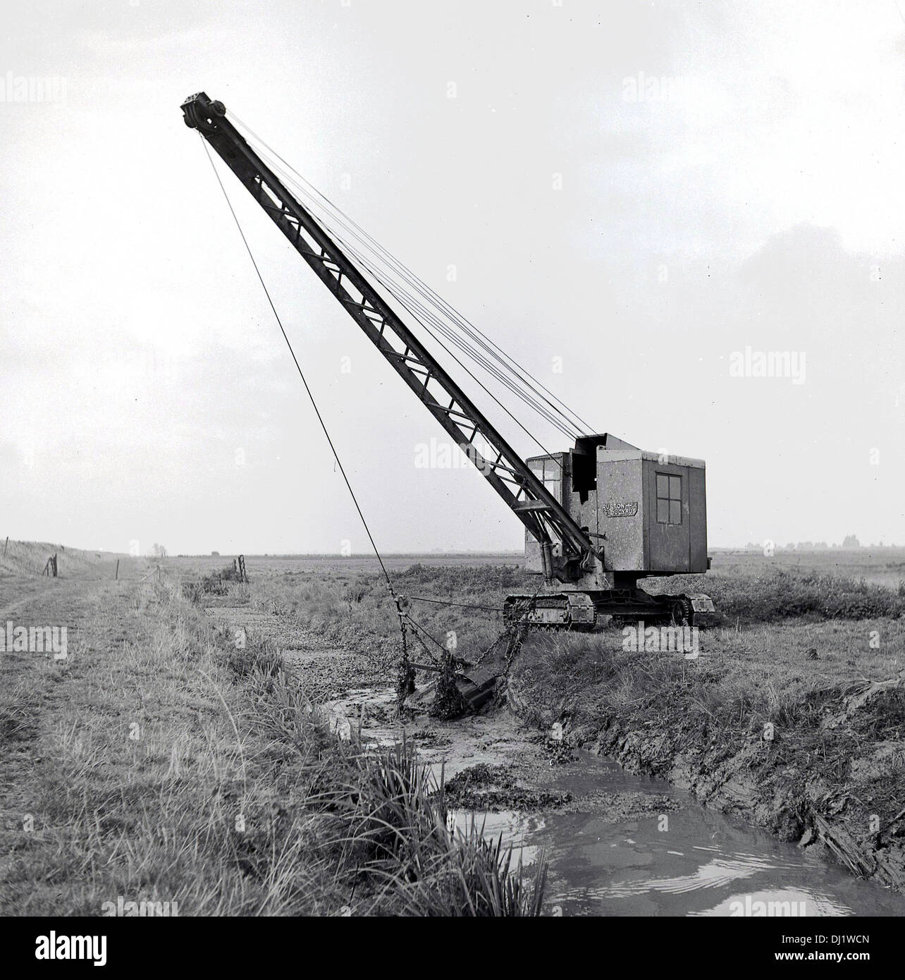 1950s, historical picture of a mechanical dragline bucket on crane with tracks positioned in a country field, excavating earth or mud from a small river or brook that runs beside it, England, UK. Stock Photo