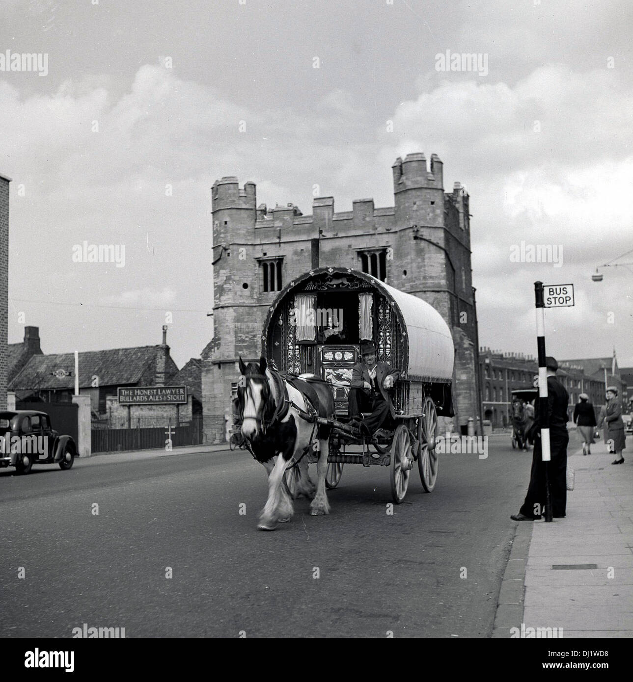 1950s historical picture of horse drawn Bow Top covered wooden wagon going pass a Bus Stop, England, UK. Stock Photo