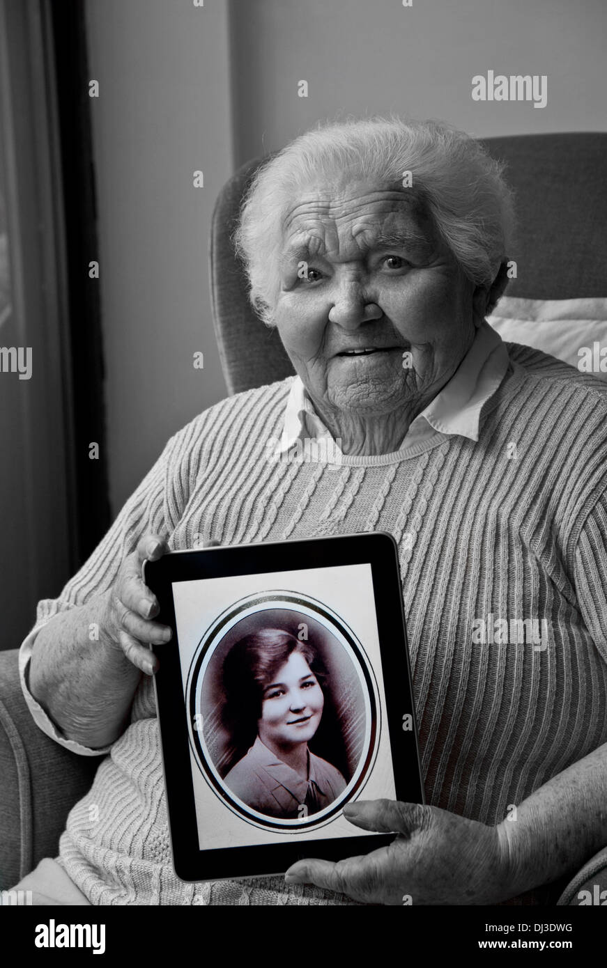 98 years old elderly lady holding an iPad tablet computer, displaying a sepia portrait of herself taken 80 years ago at age 18 Stock Photo
