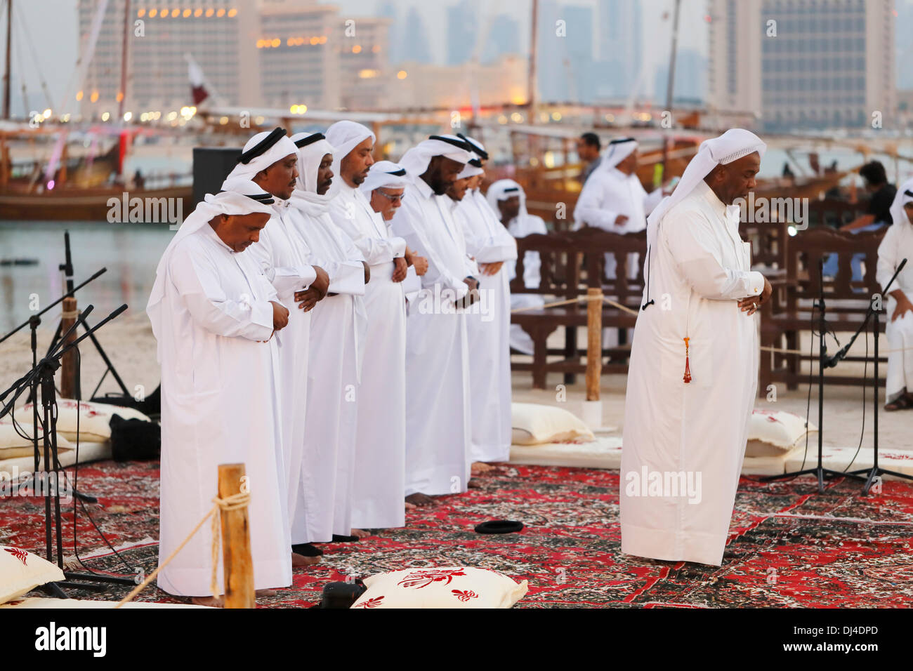 DOHA, Qatar - Nov 21 2013: Members of a Qatari traditional music group perform evening prayers at the 3rd Traditional Dhow Festival, held at Katara cultural village in the West Bay area of the Qatari capital. Some of the dhows and the city skyline are behind them. Credit:  Art of Travel/Alamy Live News Stock Photo