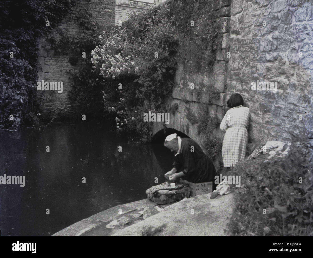 Historical picture from 1930s of an elderly lady washing clothes by a small river, with her granddaughter with her, France. Stock Photo
