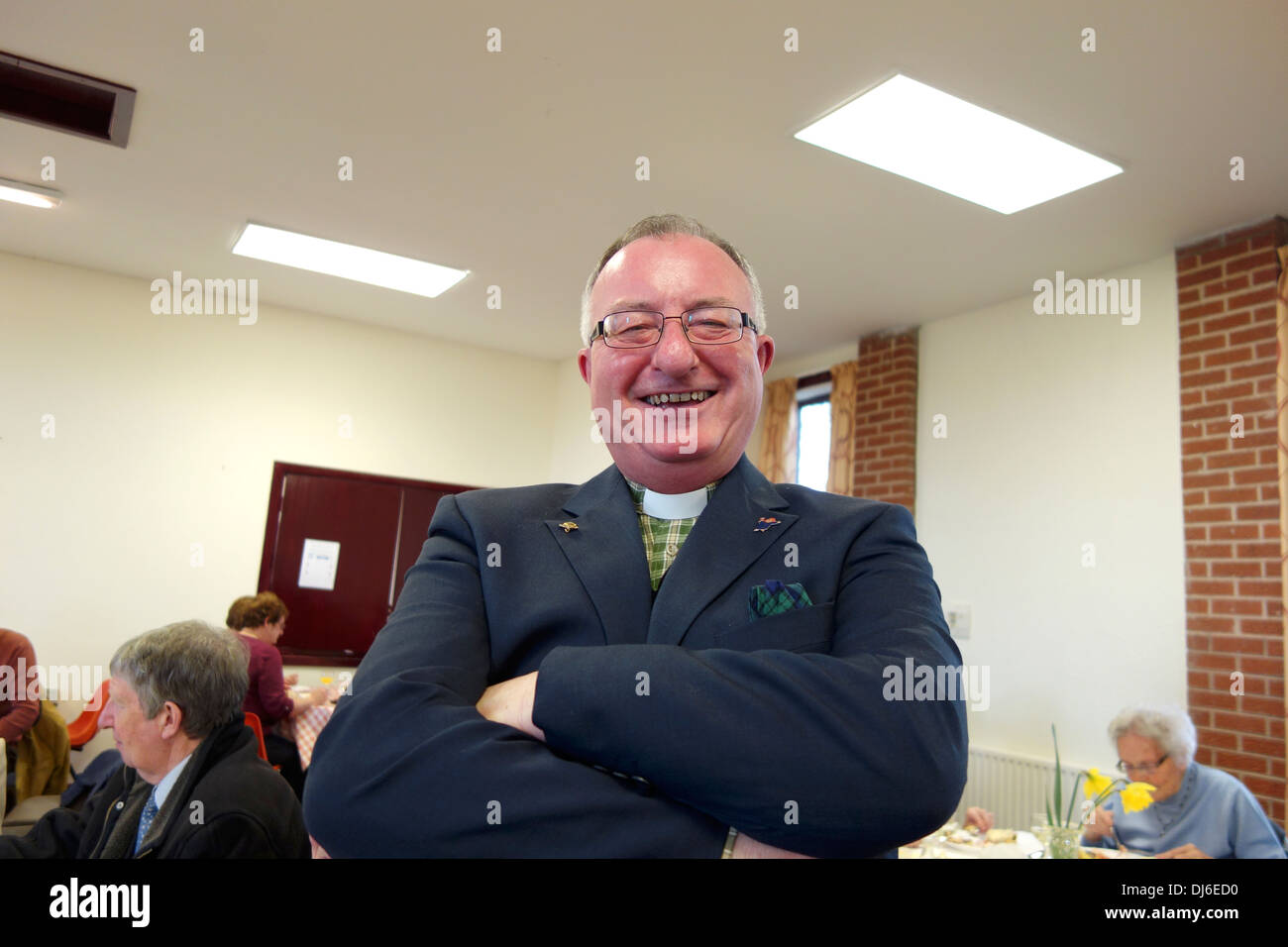 A smiling jovial looking vicar stood in a village hall with his arms folded Stock Photo