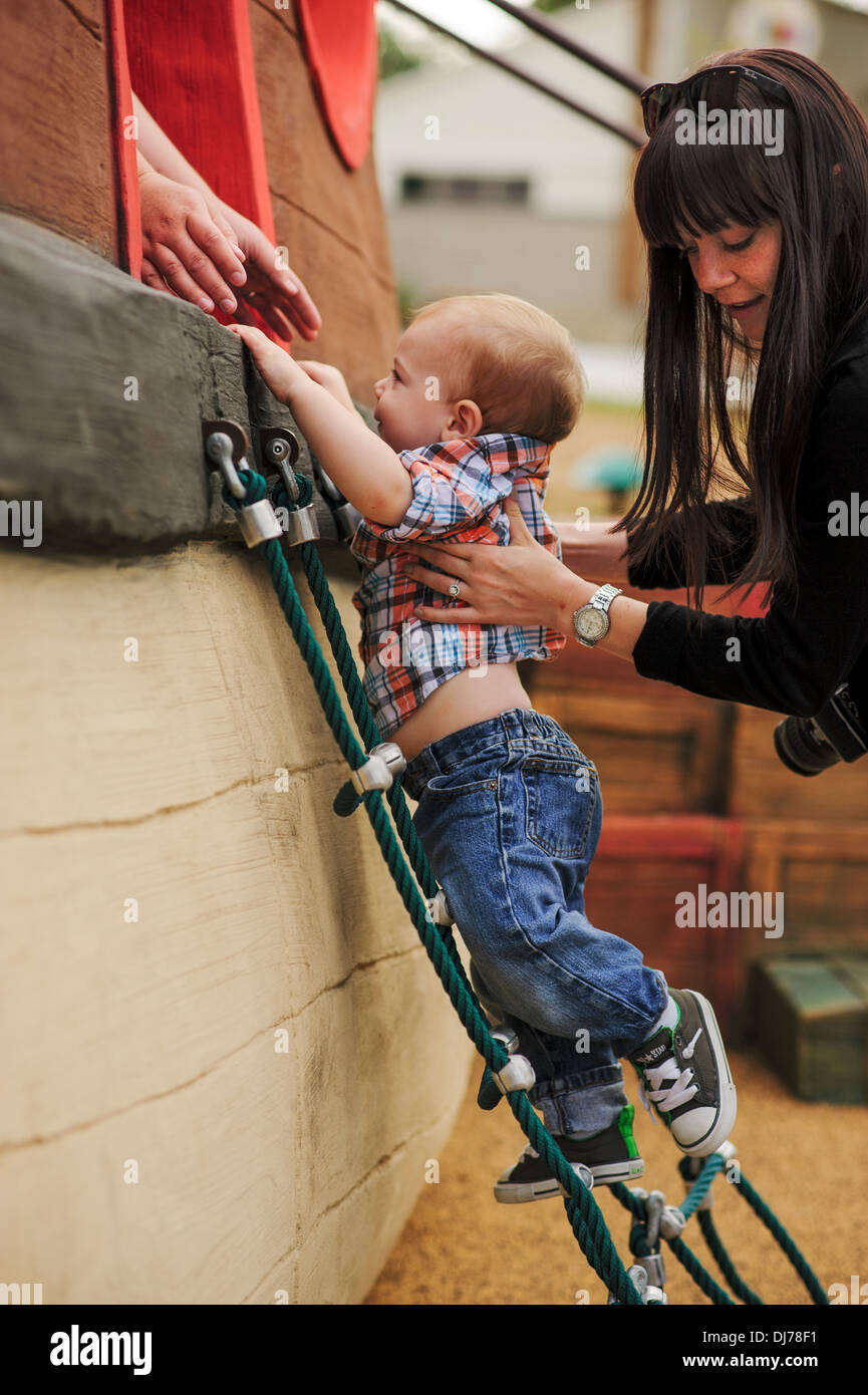 A young toddler strives to climb the ladder to reach the next level. He has the help of others. Stock Photo